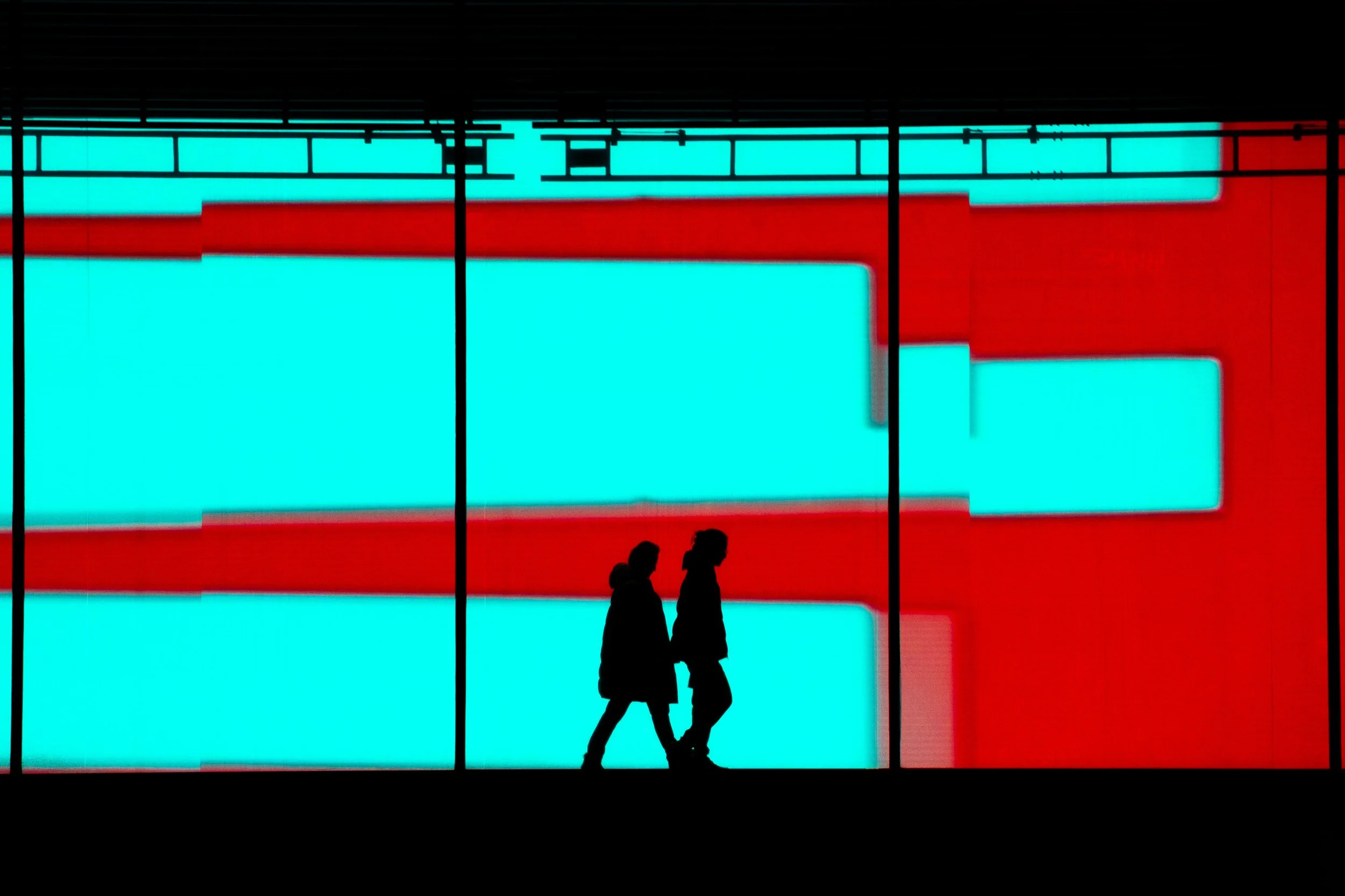 Two people walking past a brightly lit, colorful modern building with large, illuminated blue and red panels.