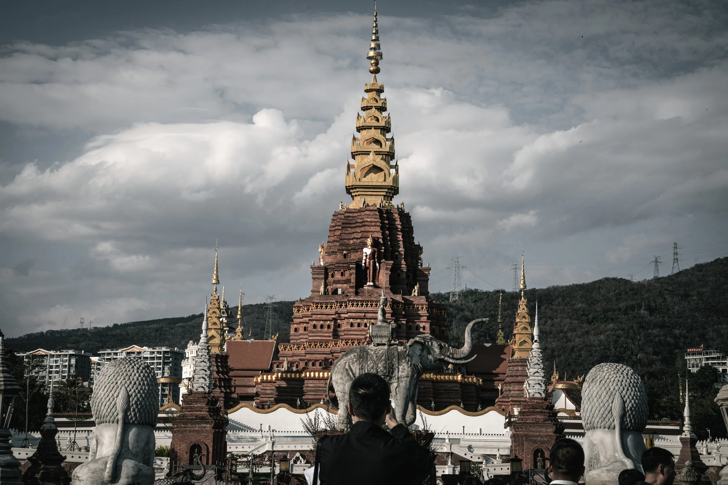 A person taking a photo of a Buddhist temple with a tall, multi-tiered pagoda, elephant statues, and ornate decoration, set against a cloudy sky and mountainous background.