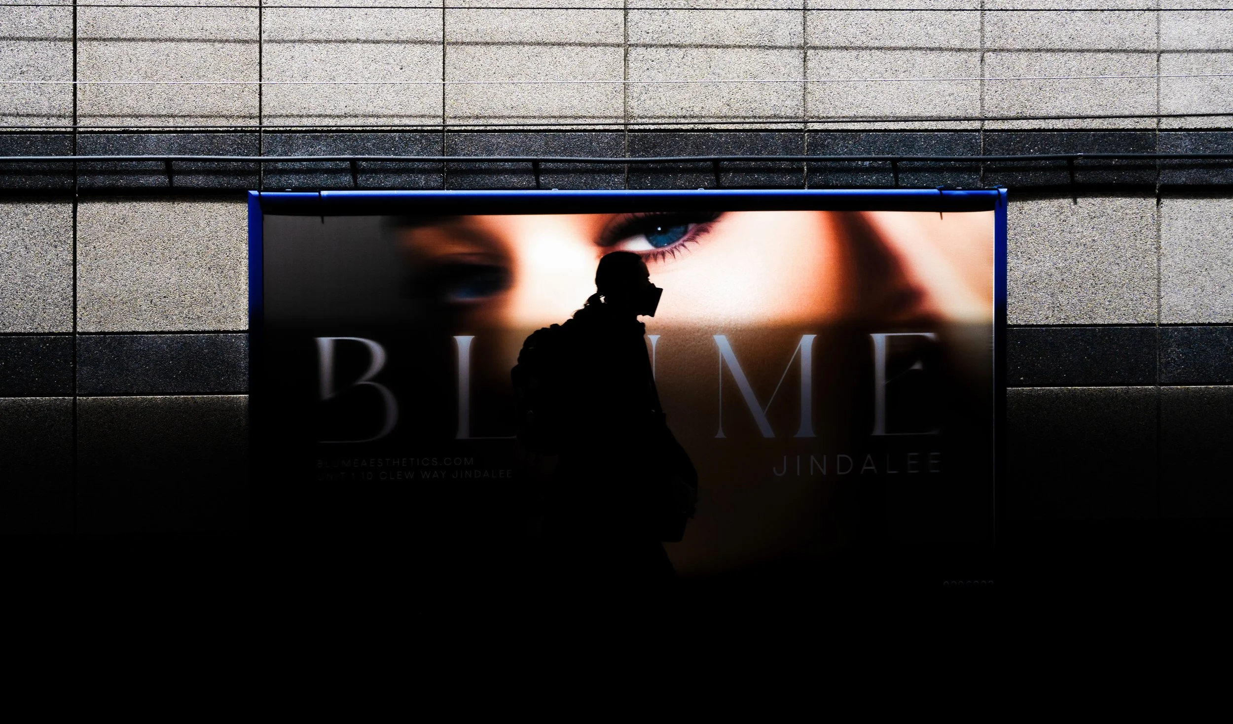 Silhouette of a person wearing a face mask standing in front of a billboard featuring a close-up of a woman's face and eye.
