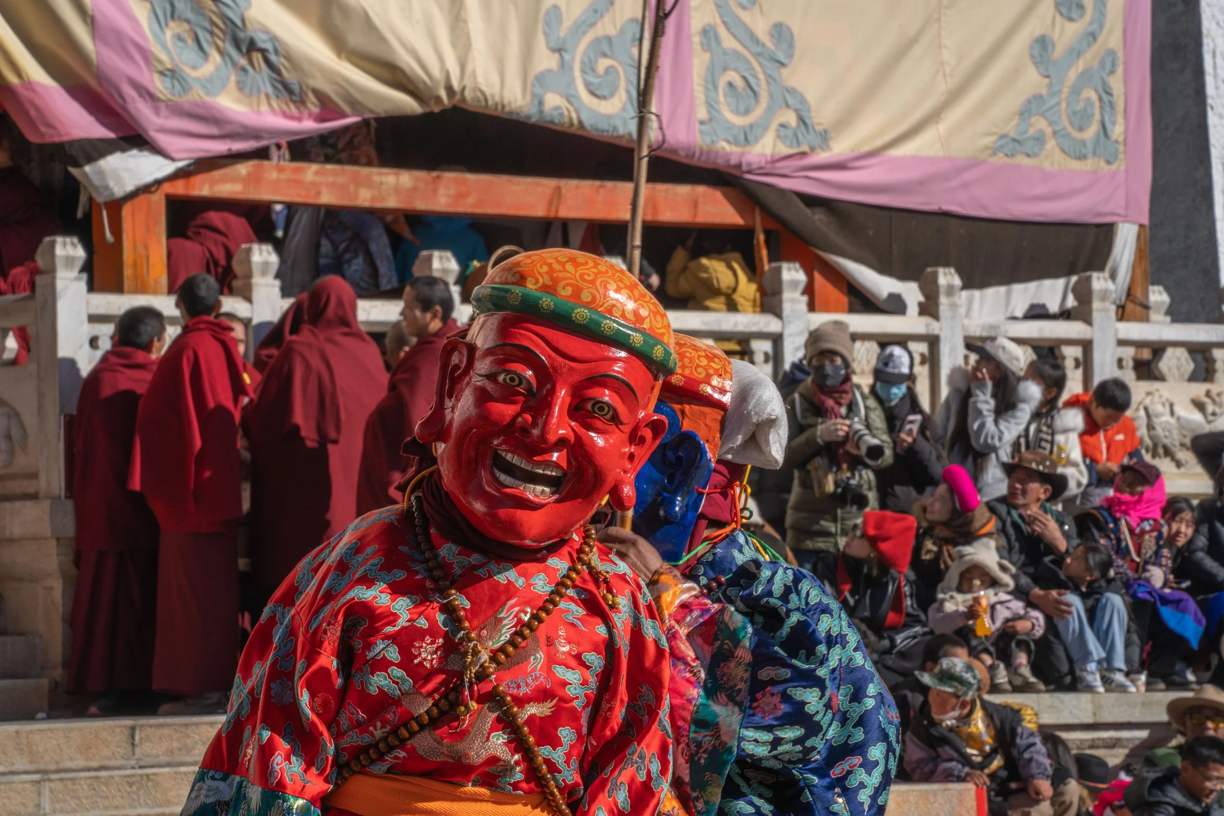 A person wearing a red costume and mask performing in a cultural event, with a crowd of spectators and monks in the background.
