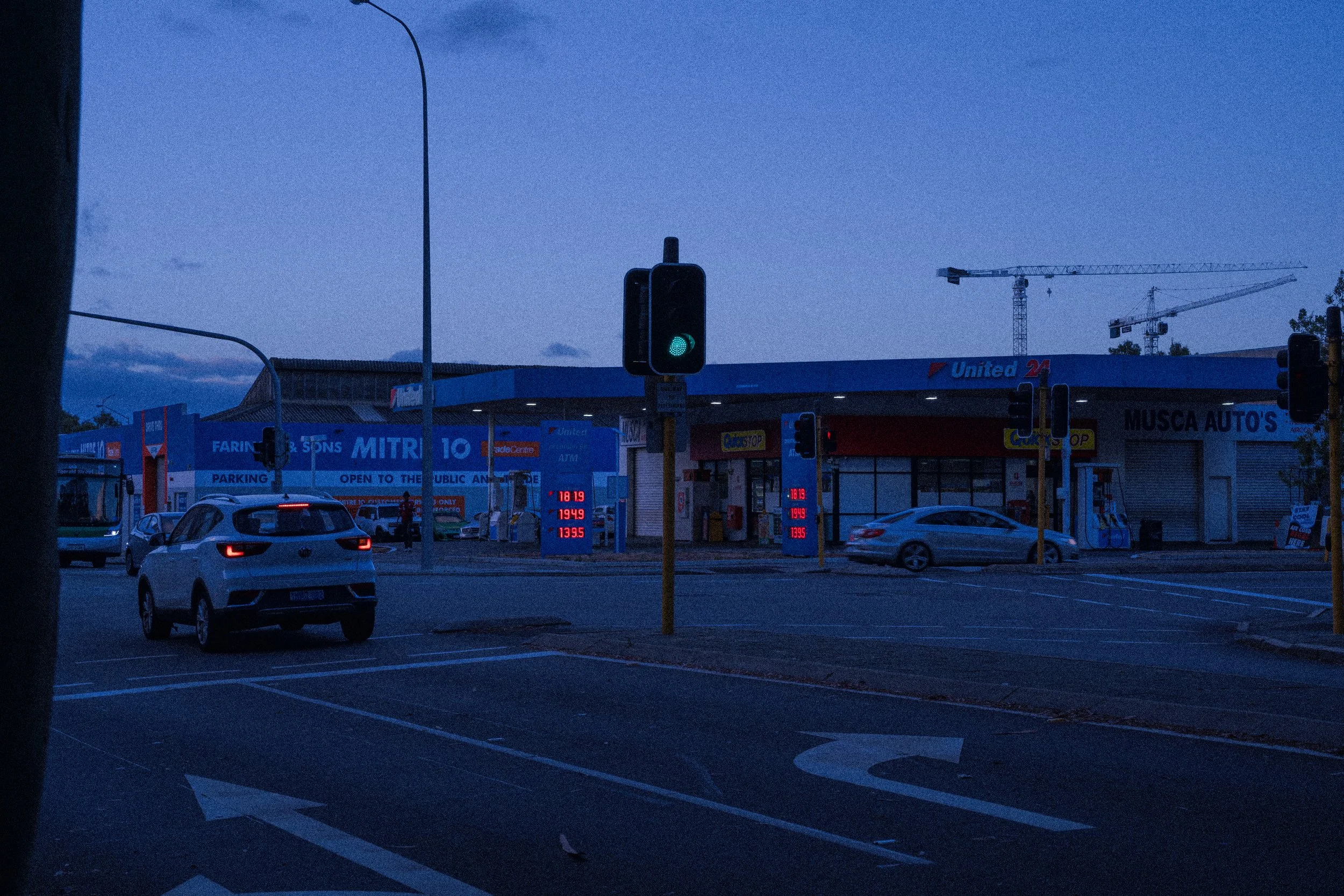 A parking lot with a green traffic light, several cars, and a building displaying fuel prices, signs, and a construction crane in the background at dusk.
