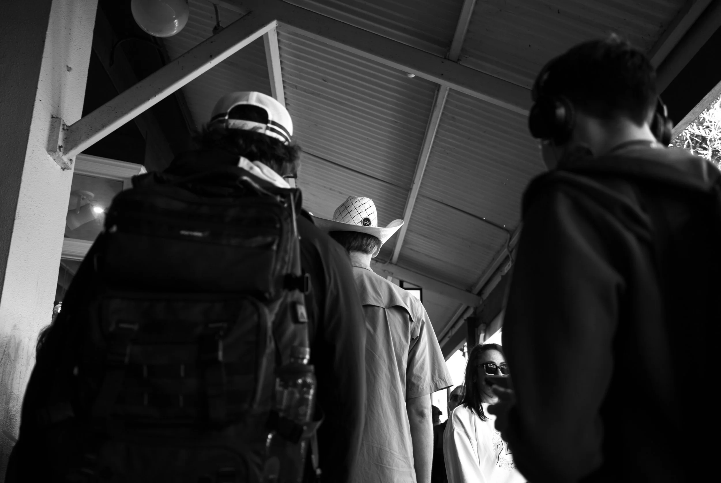 Black and white photo of several people standing outdoors under a metal-roofed structure, seen from behind, including a person with a cowboy hat and a woman wearing sunglasses.