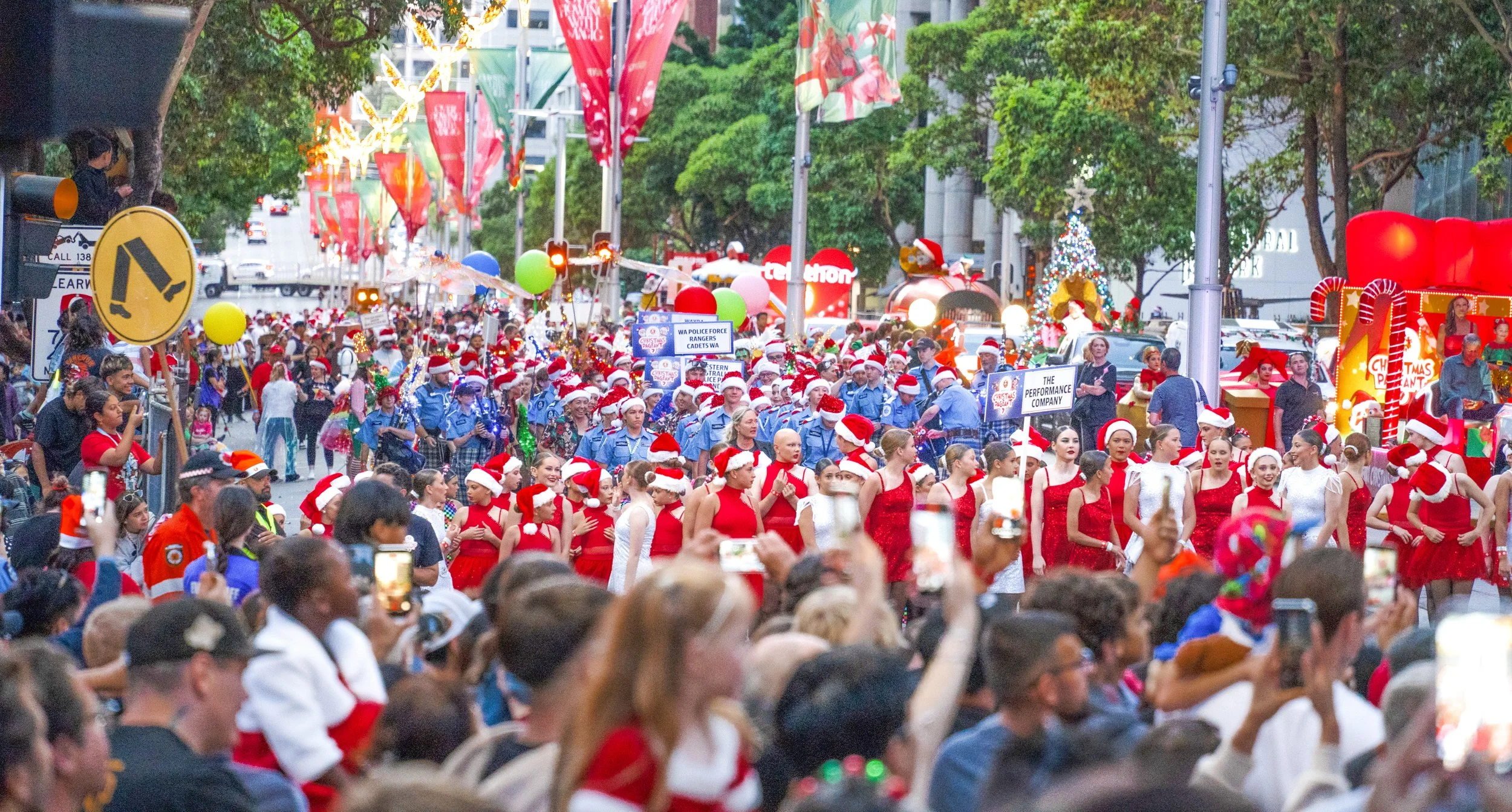 A Christmas parade with dancers dressed in red and white, many wearing Santa hats, walking down the street. Behind them, a large crowd of spectators, some taking photos, and a float decorated with Christmas themes and candy canes are visible. The str