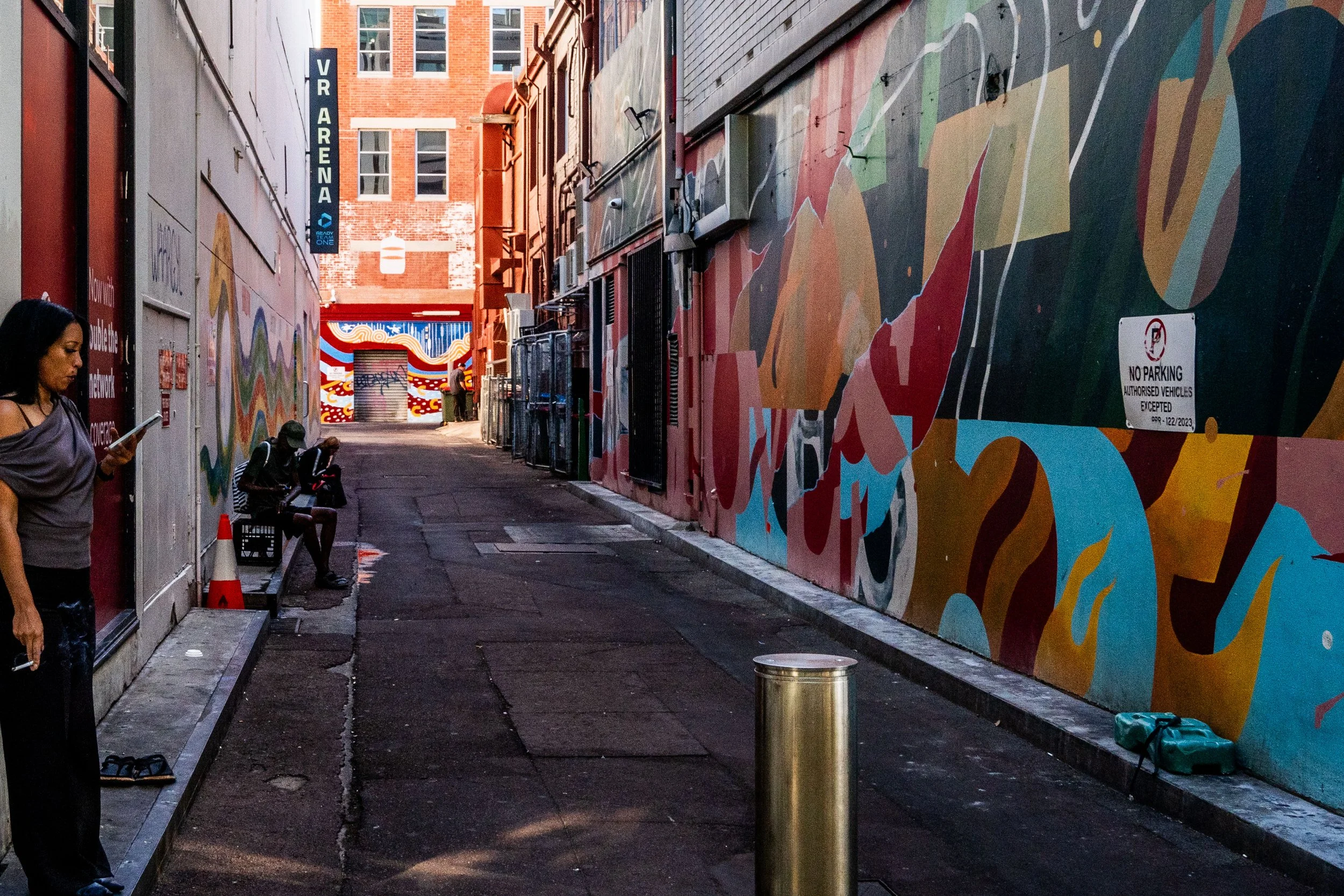 An alleyway with colorful murals on the wall, two women seated and another woman standing while using her phone, a bench, traffic cone, and a no parking sign.