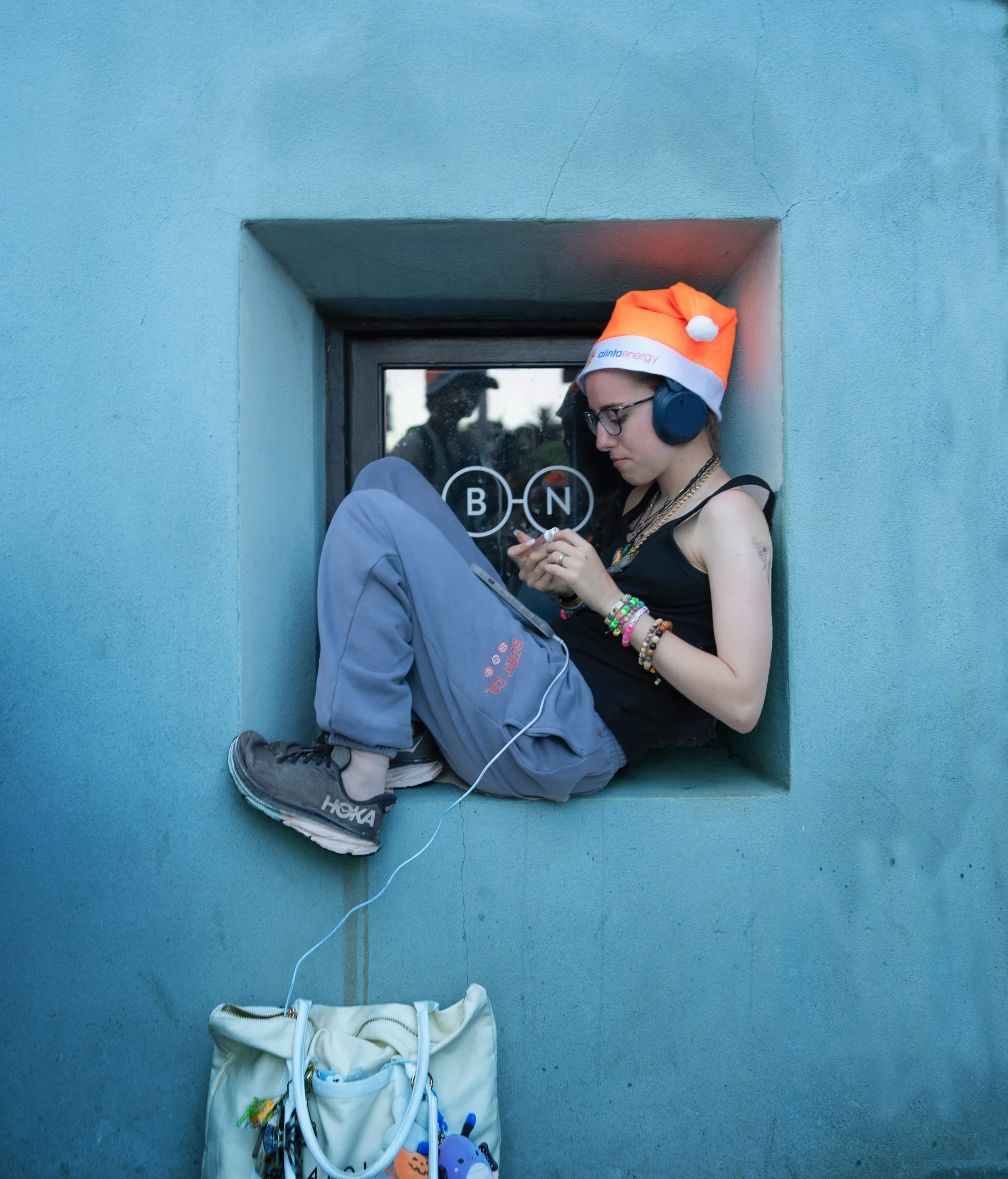 Person sitting in a small window sill, wearing a Christmas hat, headphones, glasses, and casual clothes, looking at a phone, with a backpack hanging below.