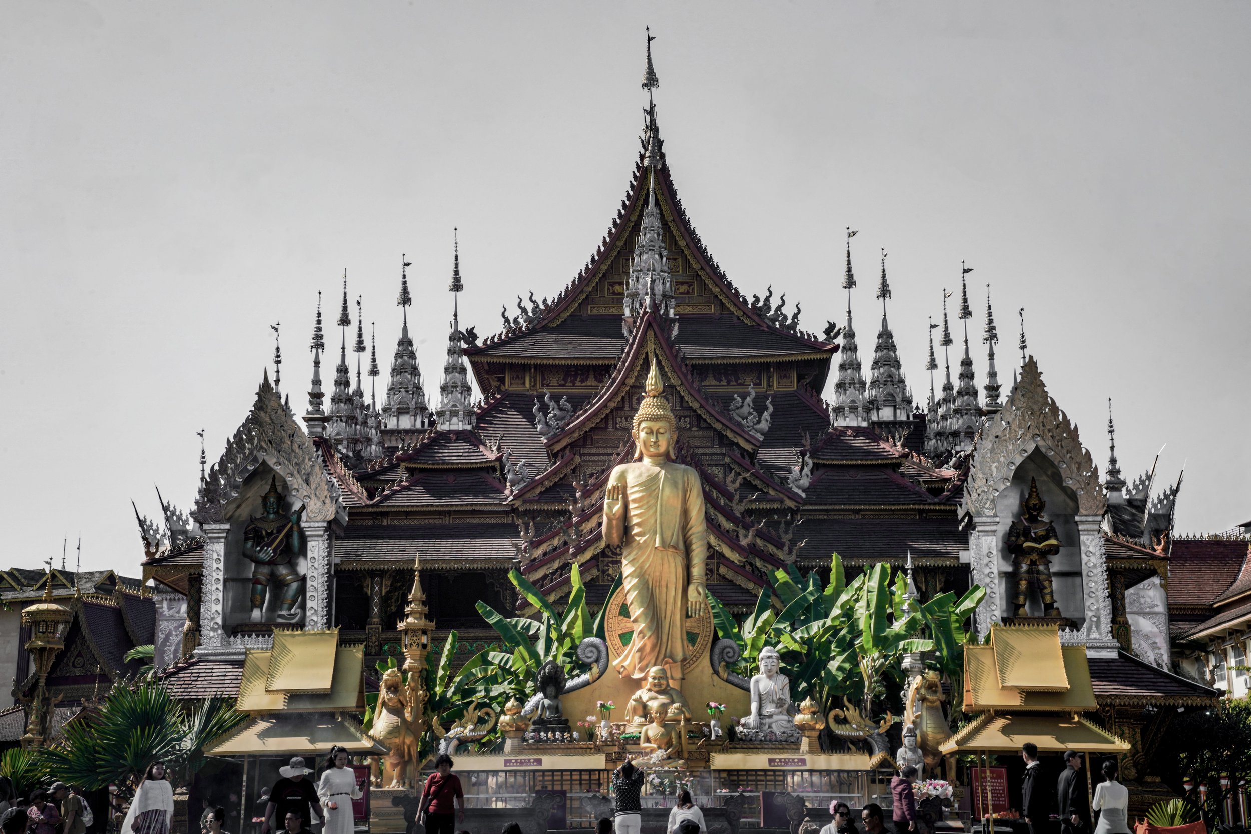 Ornate temple with multiple spires, large golden Buddha statue in front, and smaller statues, surrounded by lush green plants and visitors.