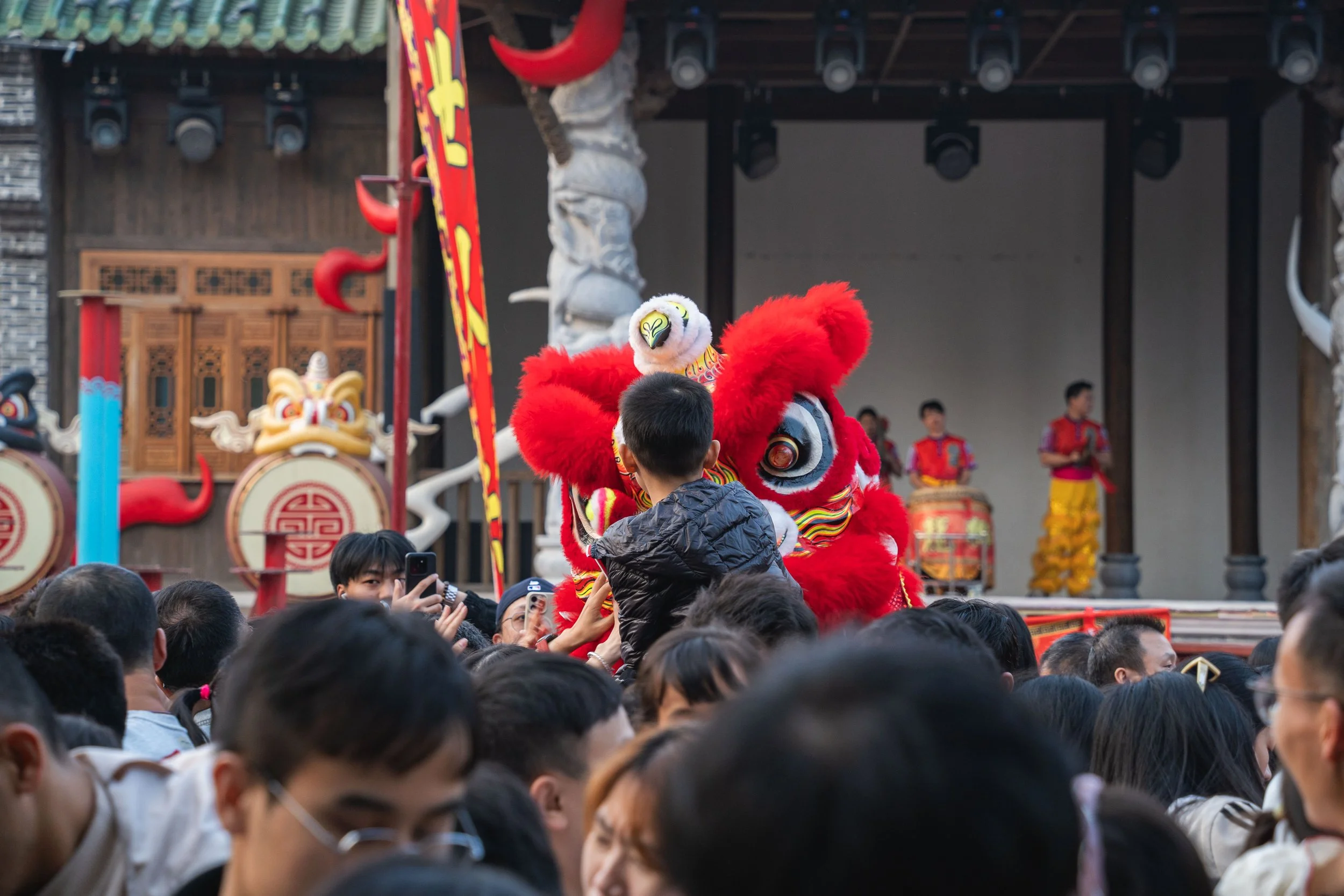 A traditional lion dance performance during a festive event with a crowd of spectators. The lion costume is red with colorful details, and performers control its movements. Behind, a stage features musicians playing drums, and decorative dragon eleme