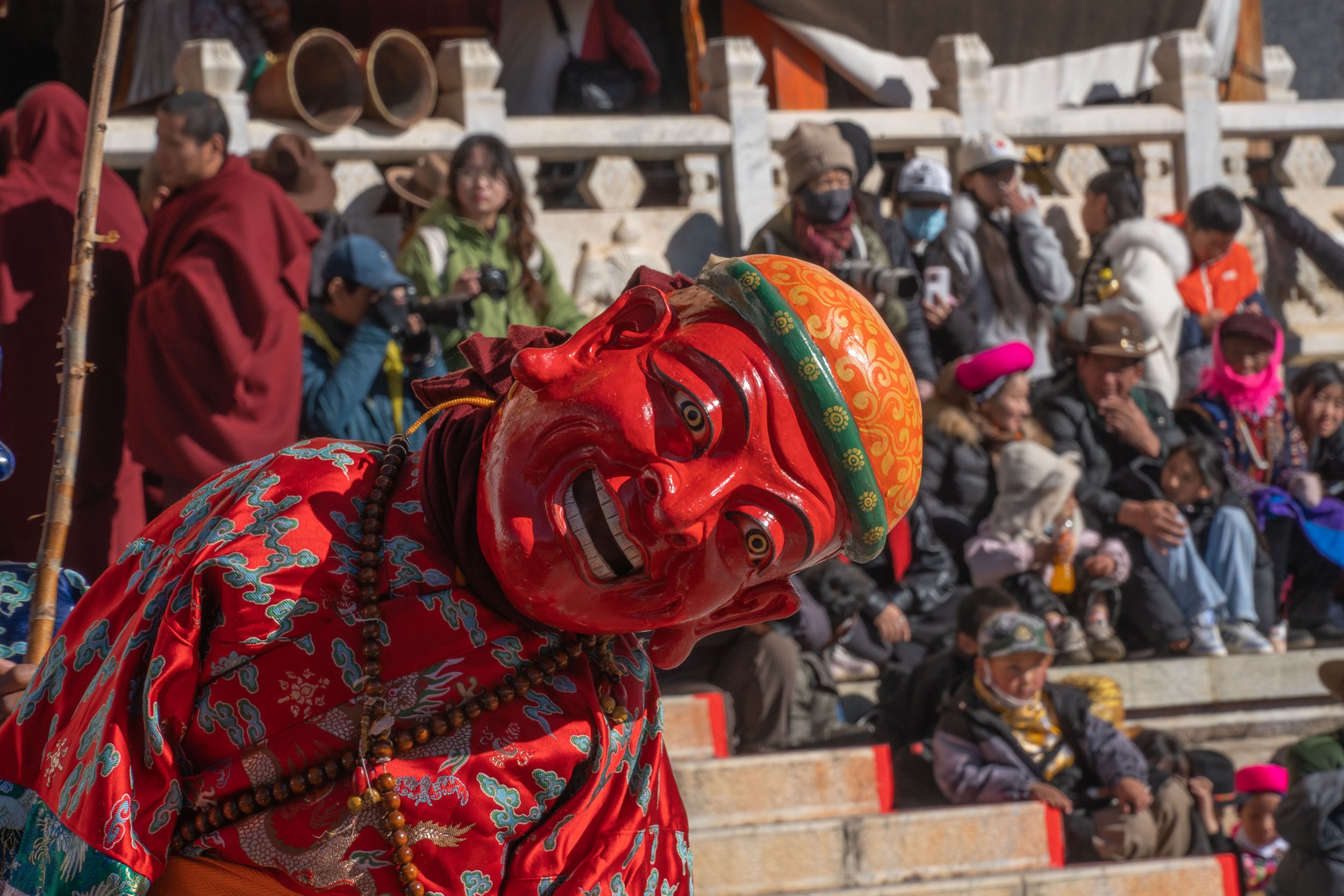 A person in a traditional red costume and a large red mask performing at a cultural event, with an audience of spectators seated in the background.