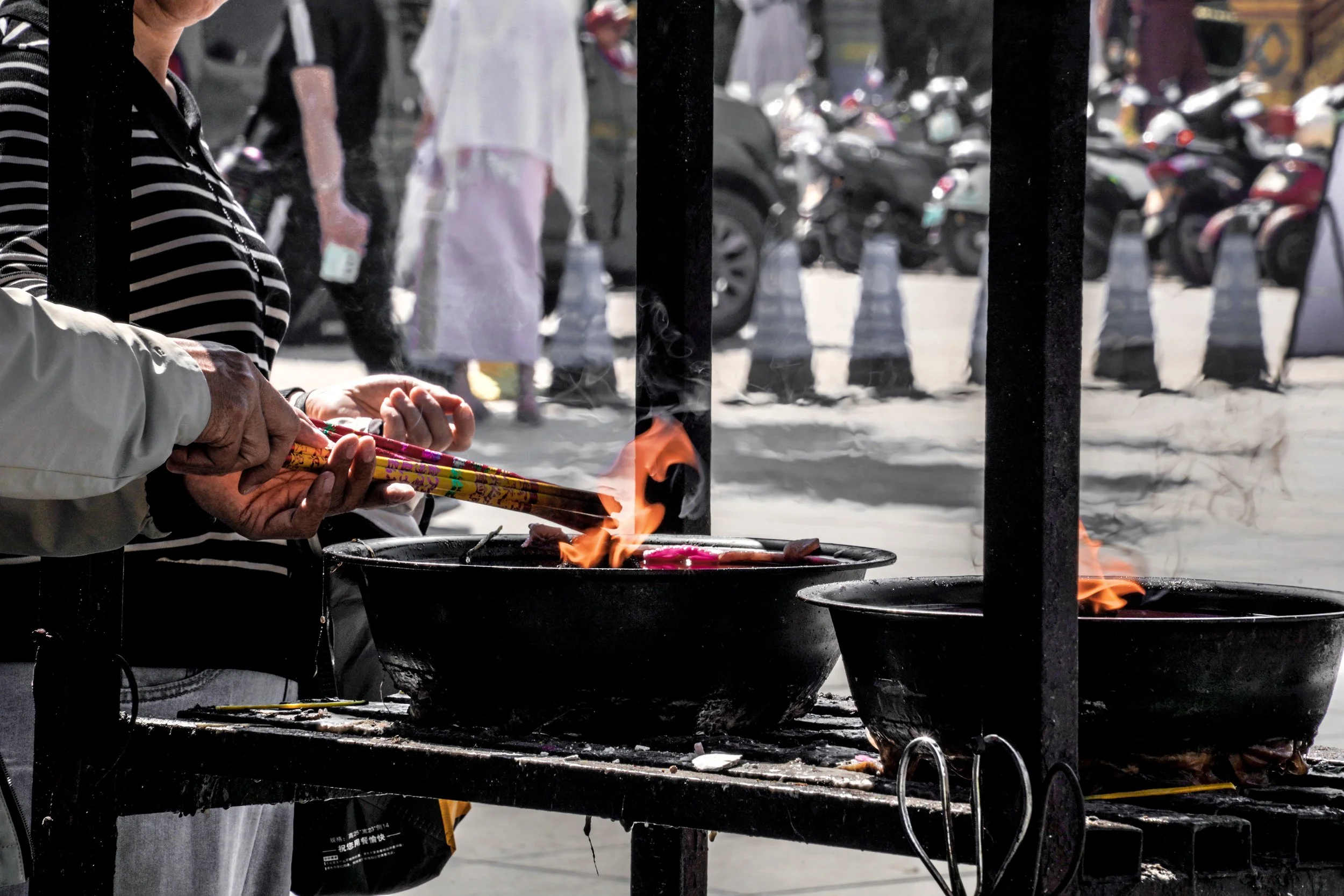 Person lighting a fire near bowls on a street food stall, with people and parked motorcycles in the background.