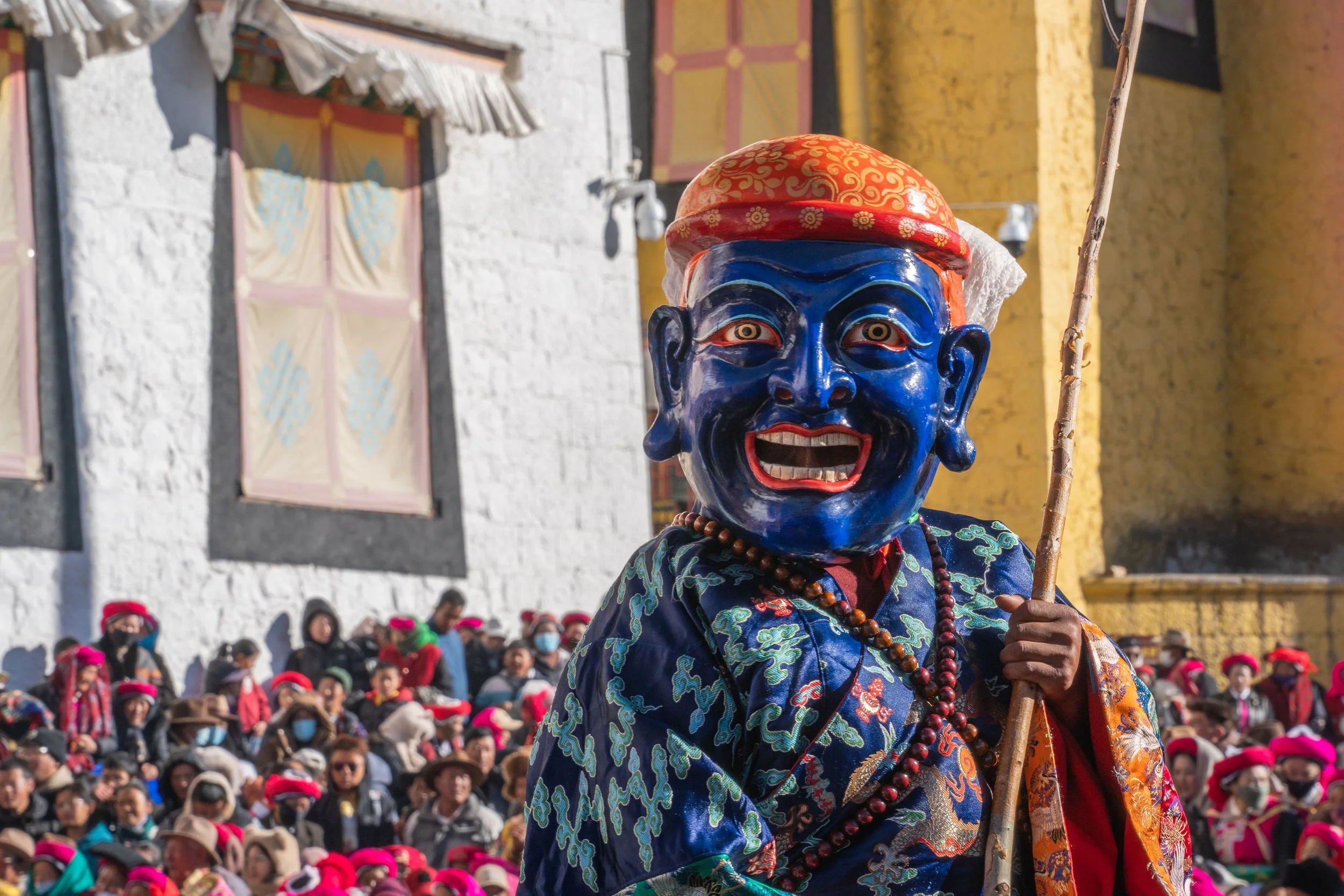 A person dressed in traditional costume with a large blue mask, red hat, and holding a stick, in front of a crowd of people wearing pink hats, outside buildings with colorful walls.