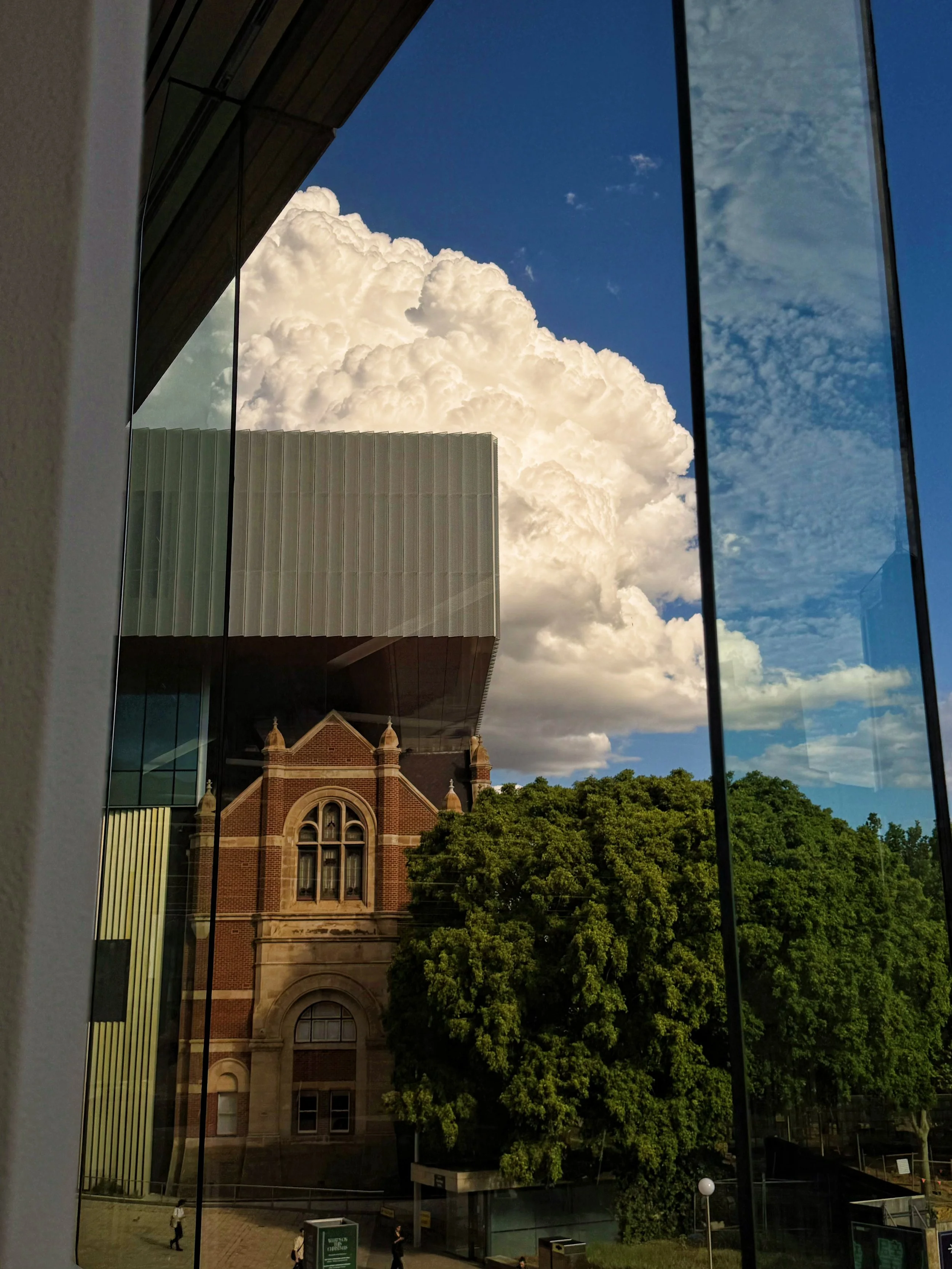 Reflections of a historic brick building with arched windows, a large green tree, cloudy sky, and modern glass building in the window reflections.