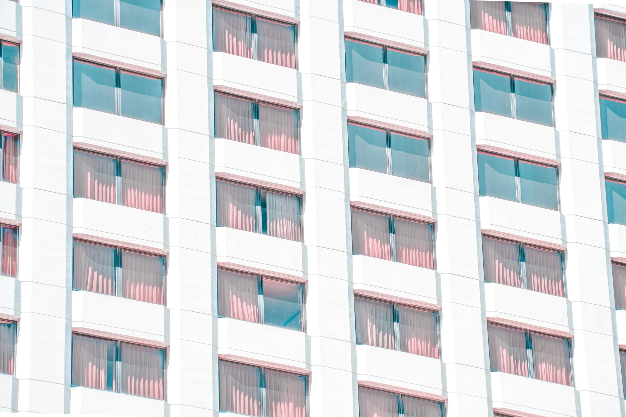 Close-up of a multi-story white building with evenly spaced rectangular windows, some with pink curtains visible behind the glass.