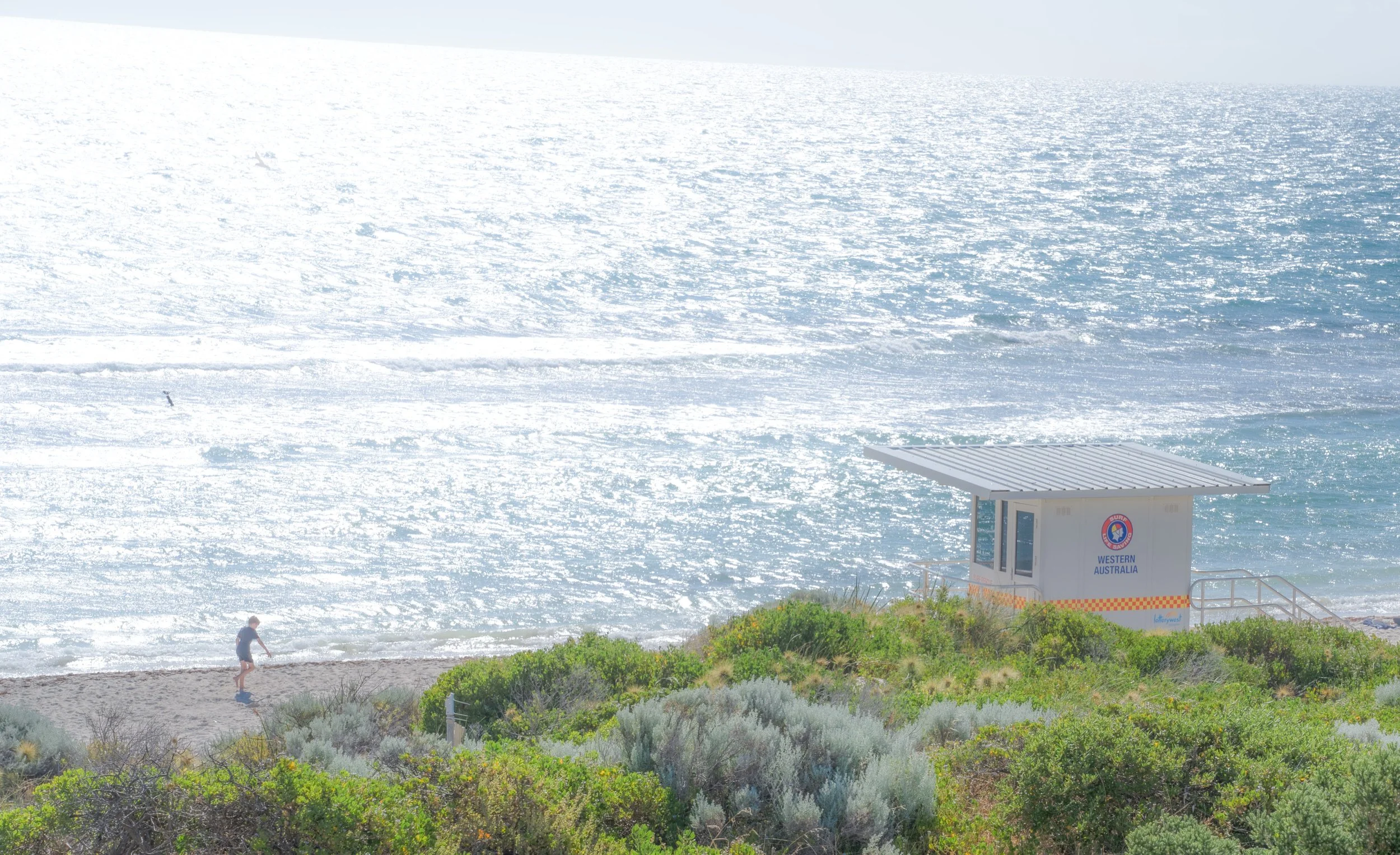 A beach scene with a lifeguard tower labeled 'Western Australia' on the right and a person walking on the sand near green shrubbery, with the ocean in the background reflecting sunlight.
