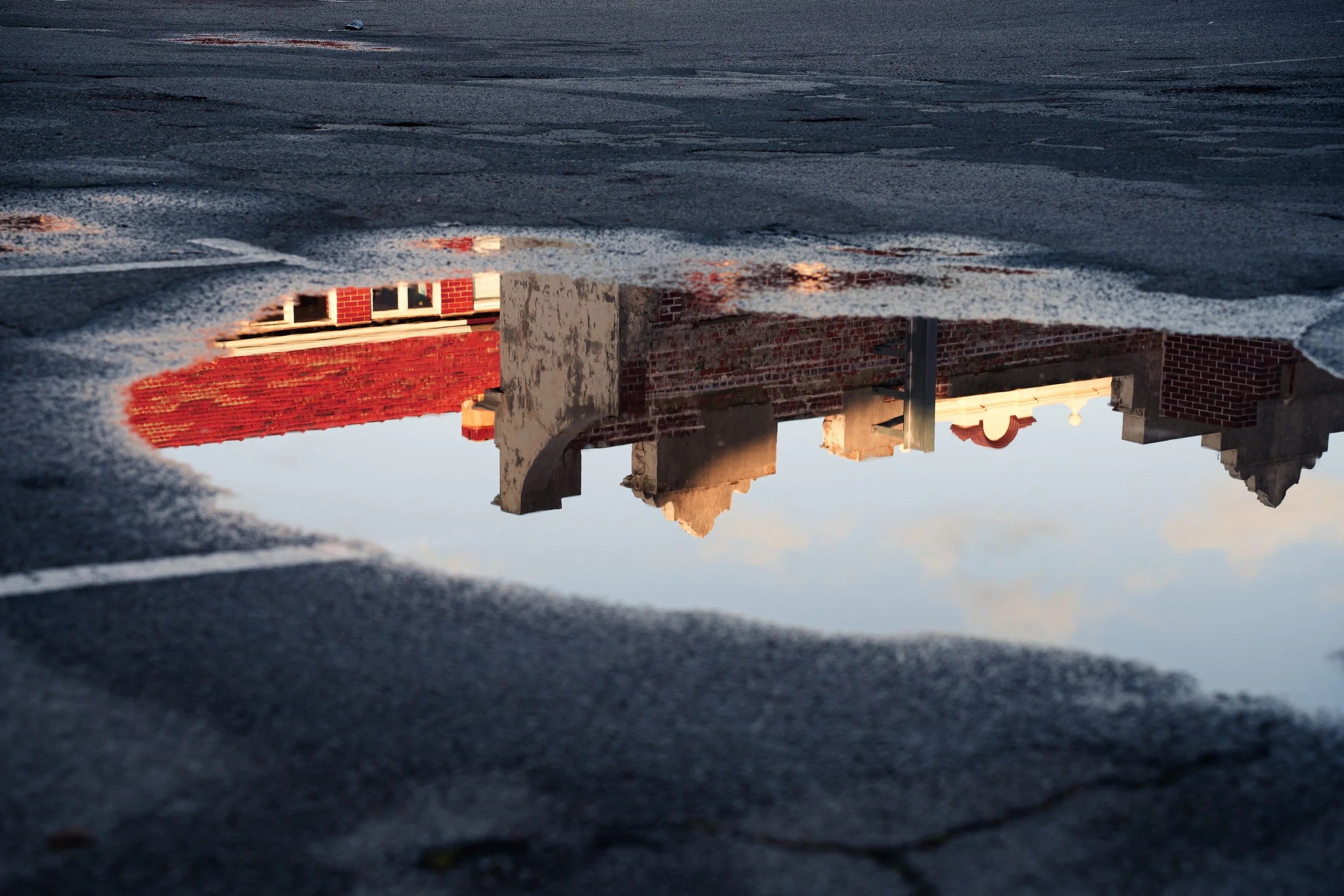 Reflection of brick buildings in a puddle on the ground, viewed upside down with the sky visible in the background.
