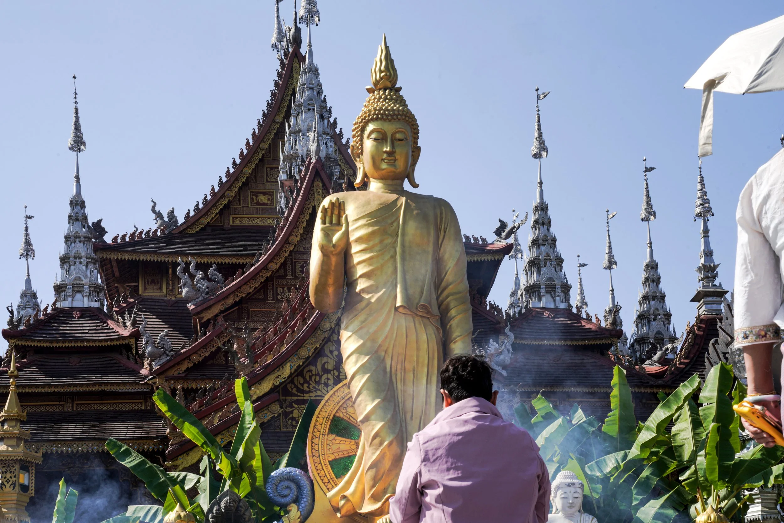 A large golden Buddha statue with an ornate temple in the background, surrounded by greenery and dedicated worshipers.
