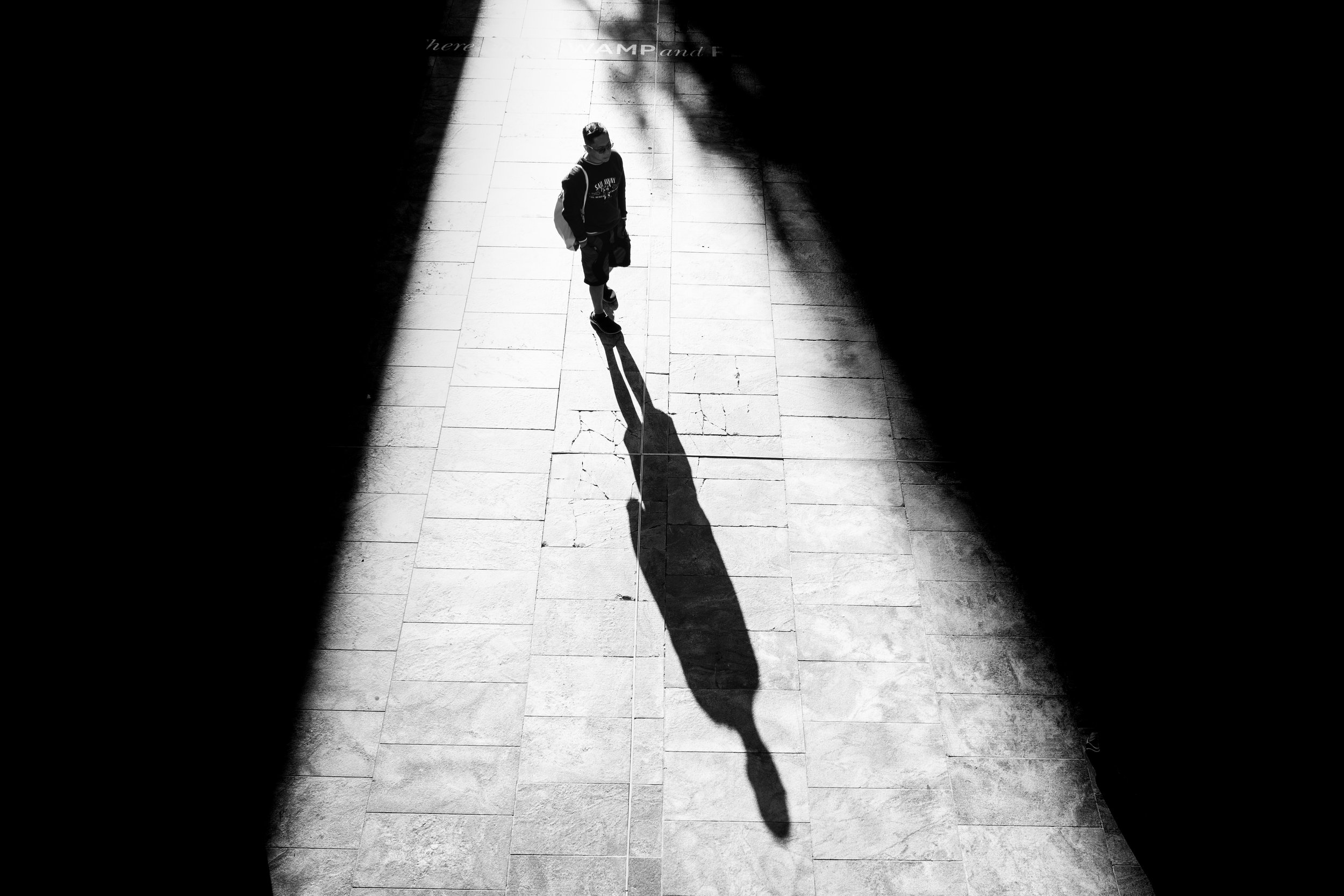 A person walking on a sidewalk with their shadow cast long and dark on the pavement, seen from above in black and white.