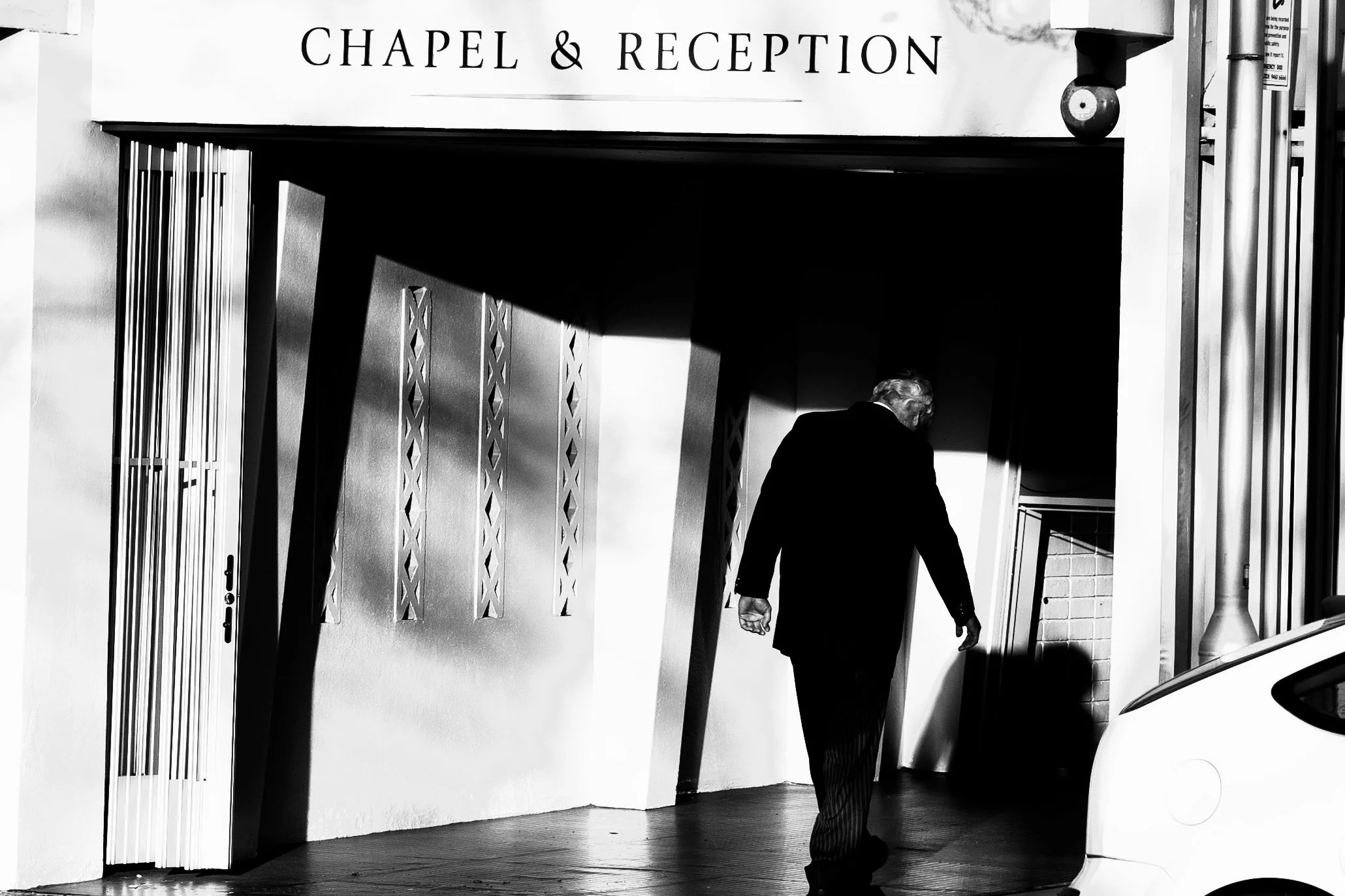 A man in a dark suit walking into a chapel and reception entrance, with sunlight casting shadows on the wall and ground, in black and white.