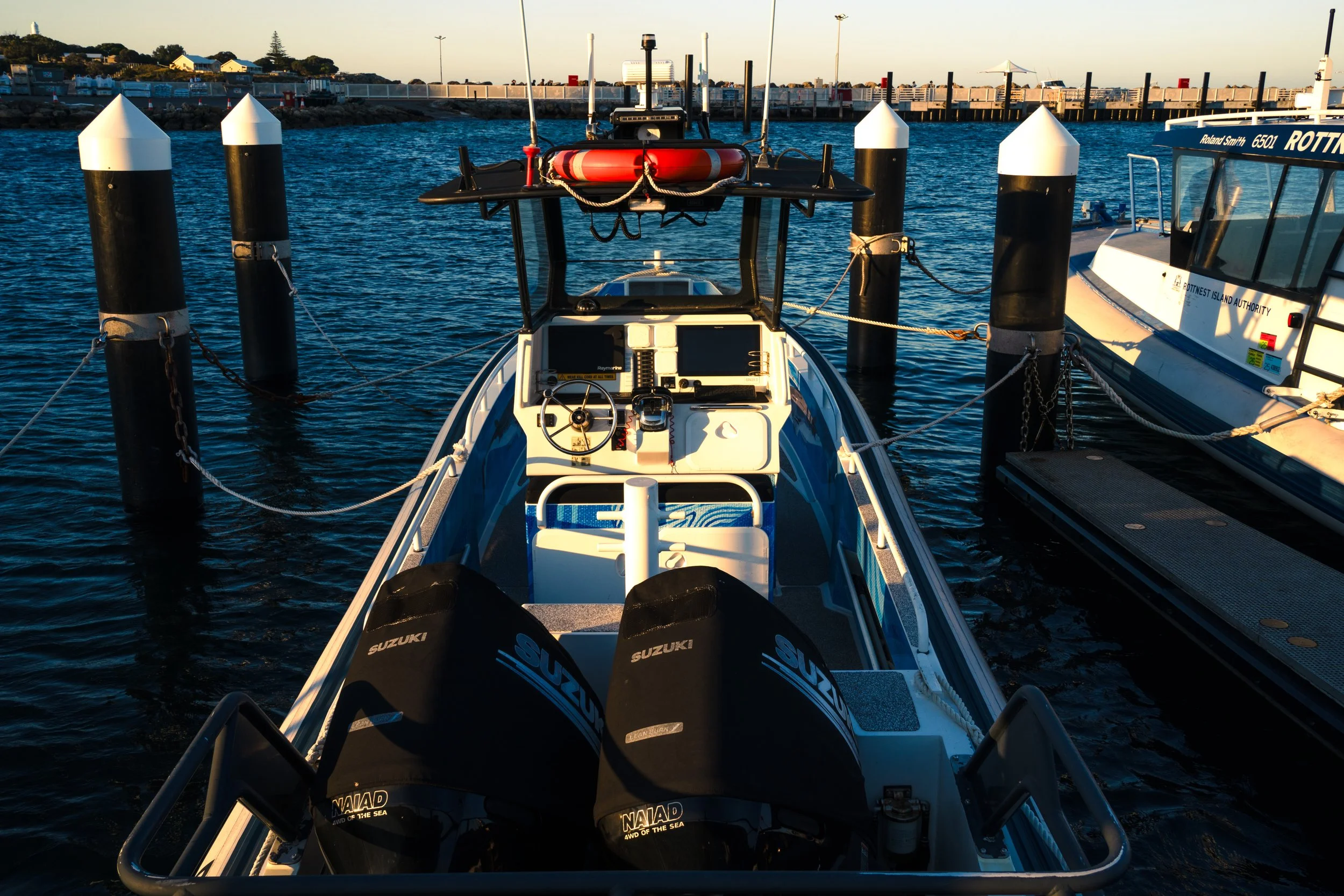 View of a small boat docked at a pier with two black outboard motors and a blue and white cabin, with calm water and other boats in the background.
