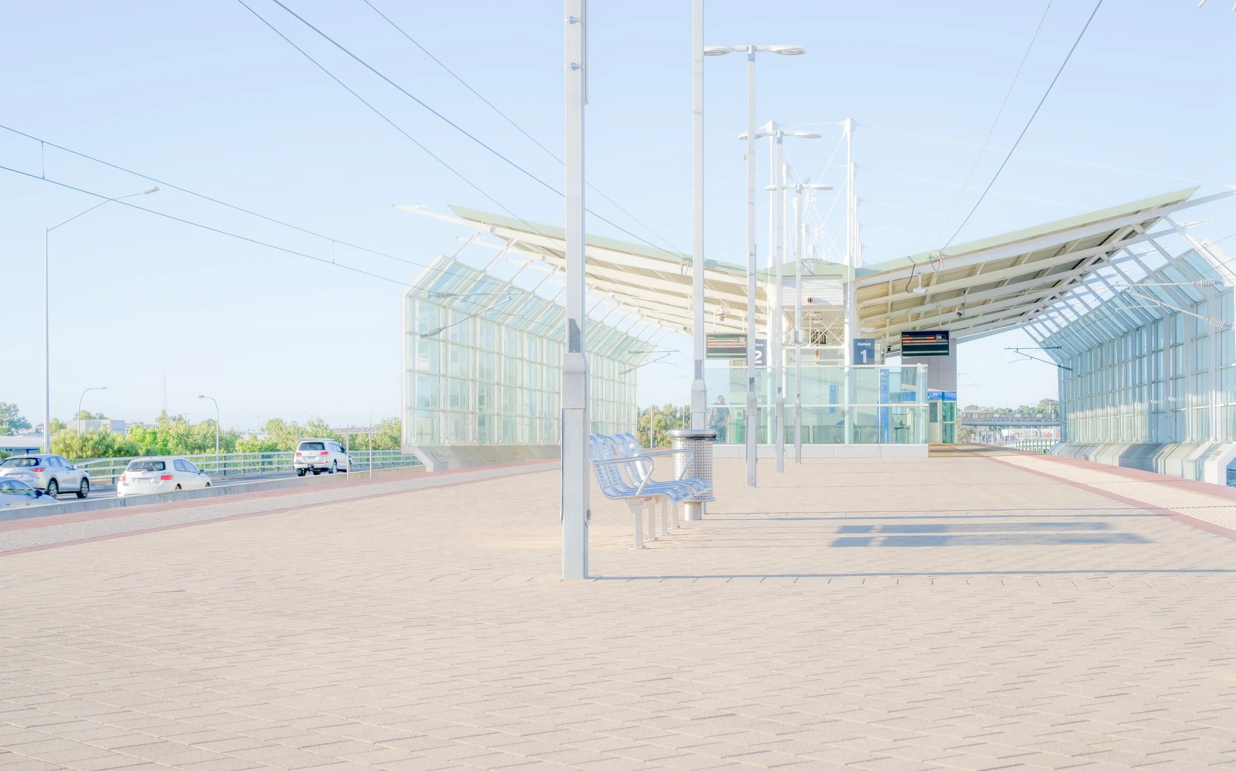 Empty bus stop with a glass shelter, benches, and lampposts, next to a road with cars, under a clear sky.