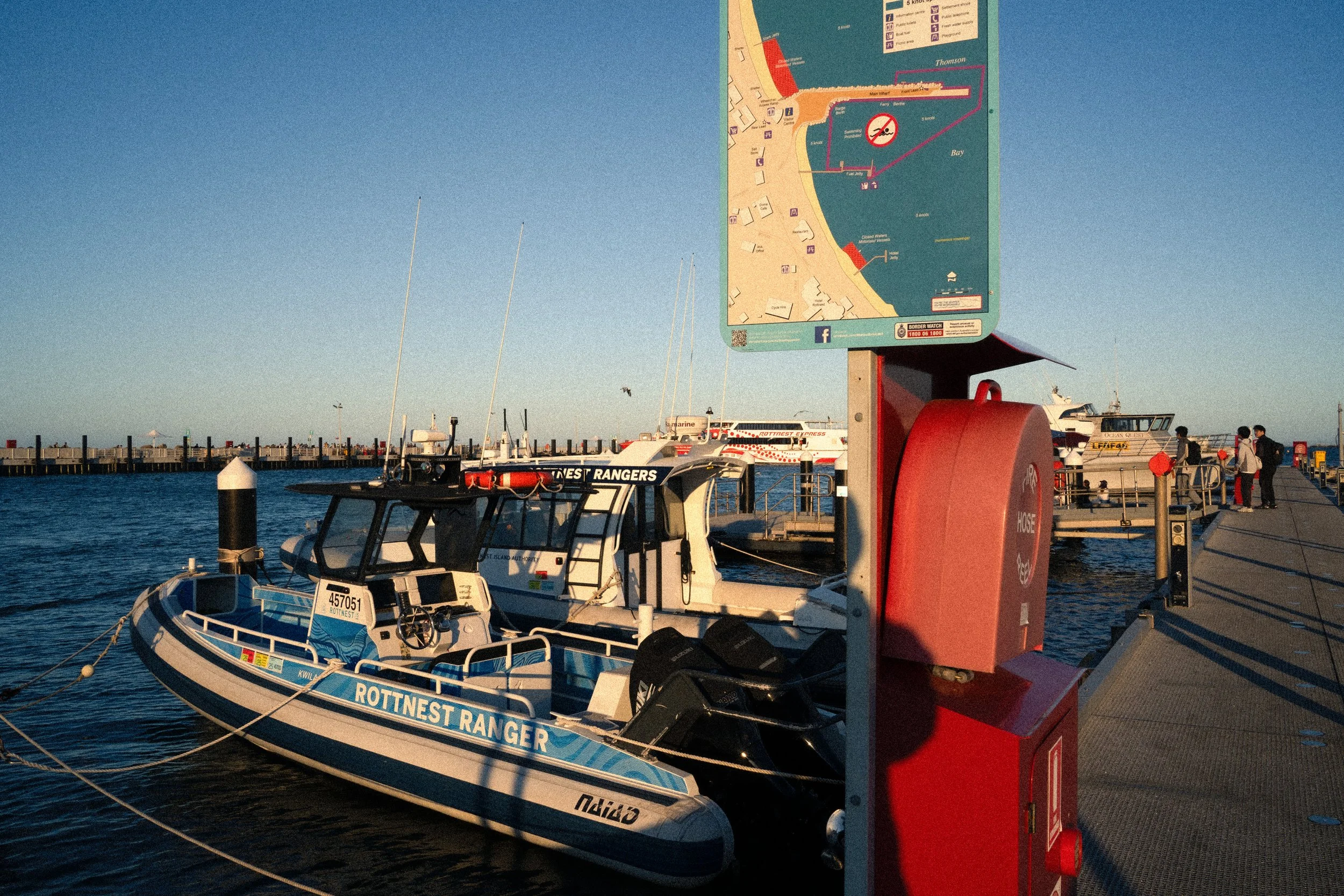 A dock with boats tied to it, including a blue and white boat labeled 'ROTTNEST RANGER'. There is a large sign with a map and a red fire hose reel on a pole at the dock.