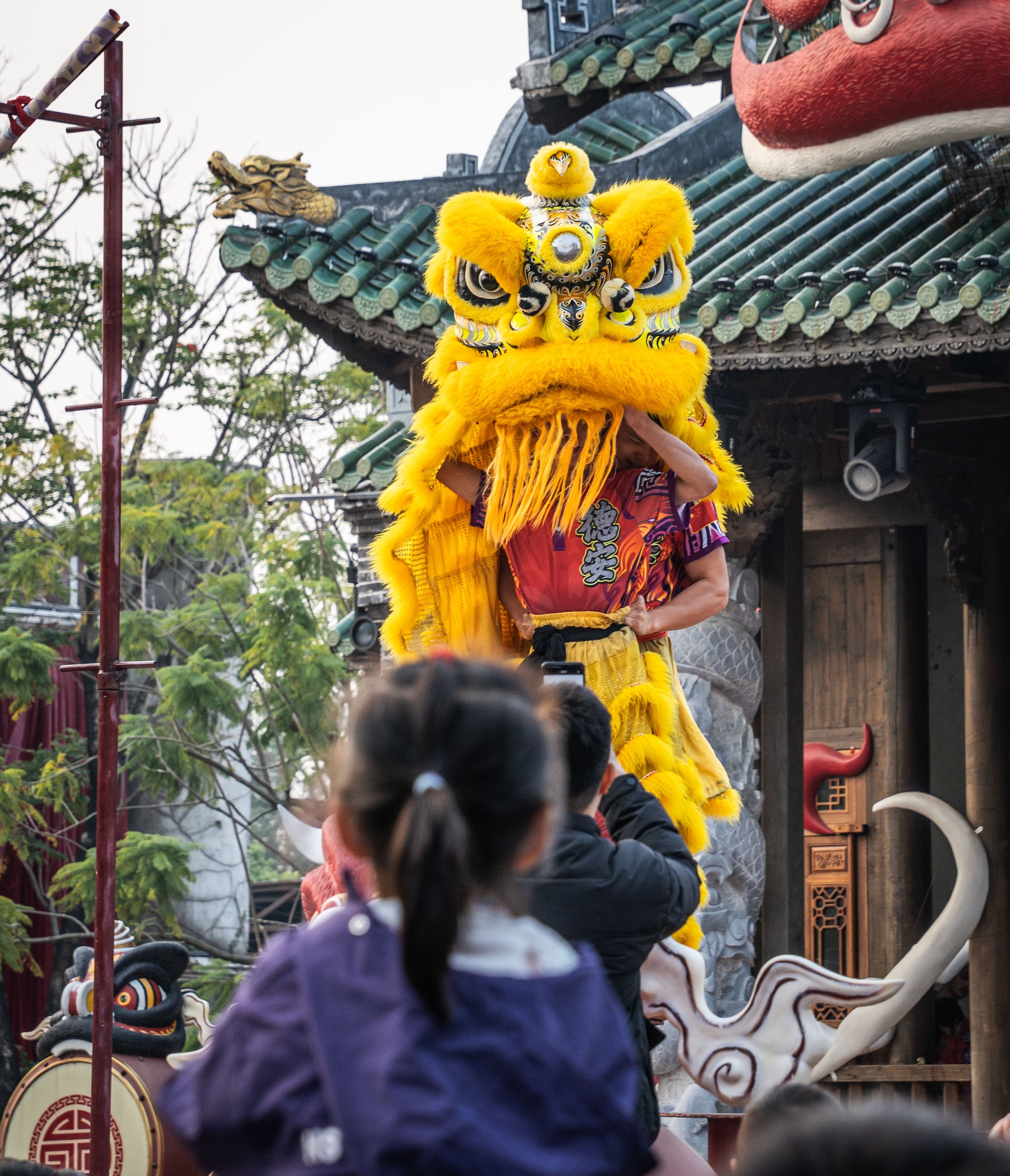 People performing a lion dance with a bright yellow lion costume on a street with traditional Chinese architecture and decorations.