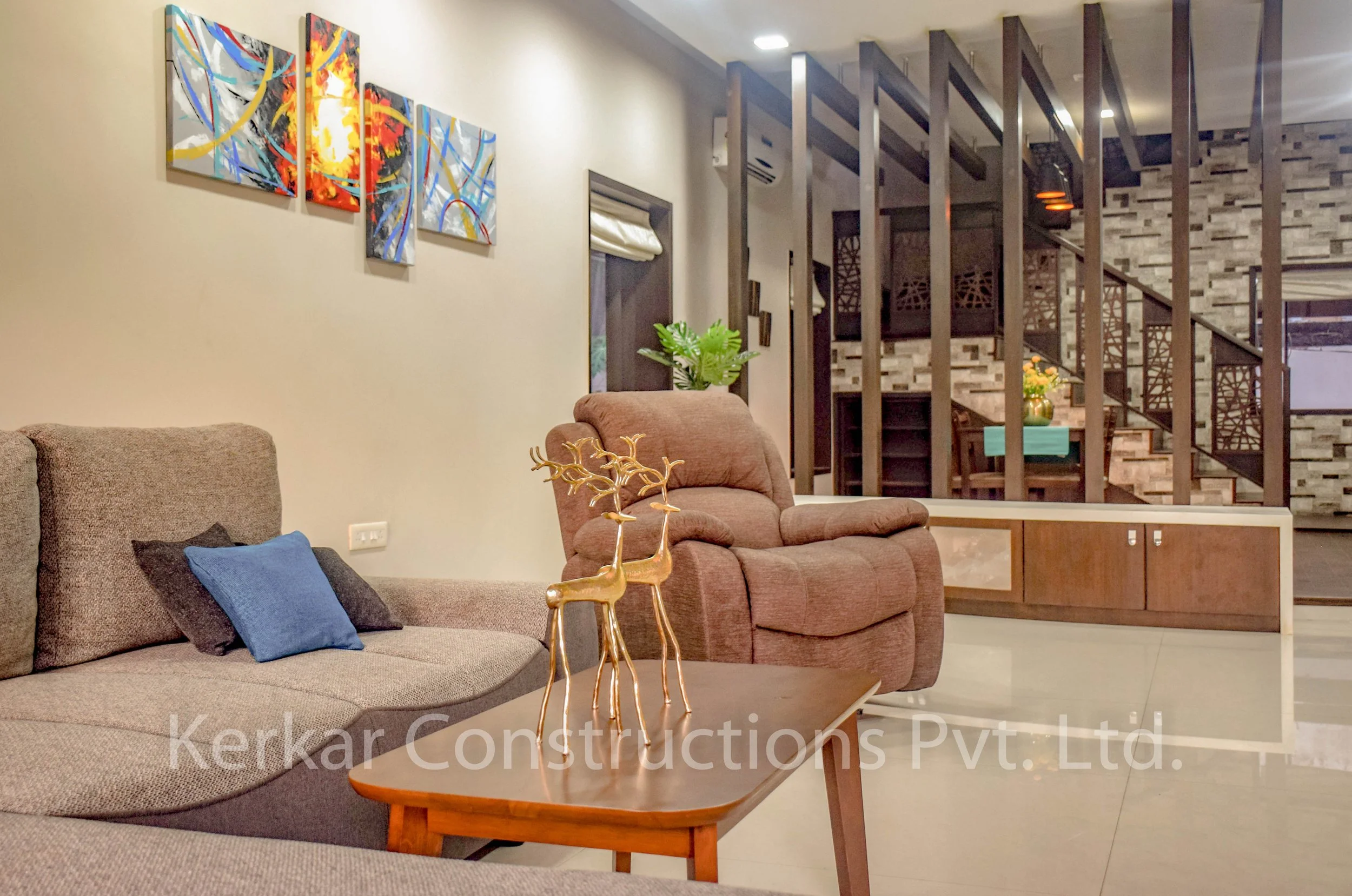 A living room with a beige sofa and a pink armchair. On the coffee table, there are two gold decorative reindeer sculptures. The wall has five colorful abstract paintings. In the background, there is a staircase with a wooden railing and a table with