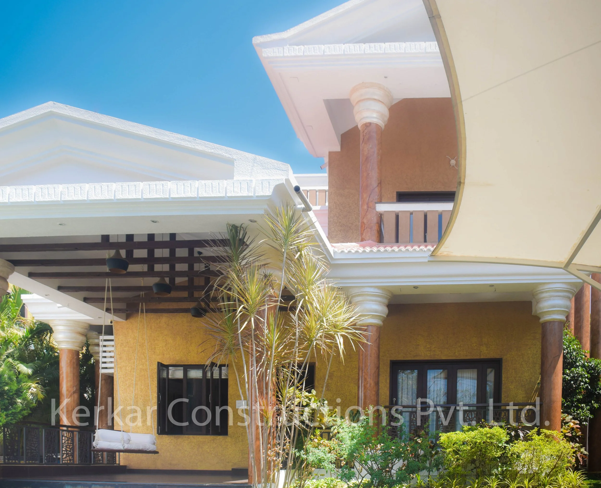 A modern house with yellow and brown exterior walls, white decorative roofing, multiple columns, and a balcony. There are lush green trees and plants in the front yard, and a swing hanging from the porch ceiling.
