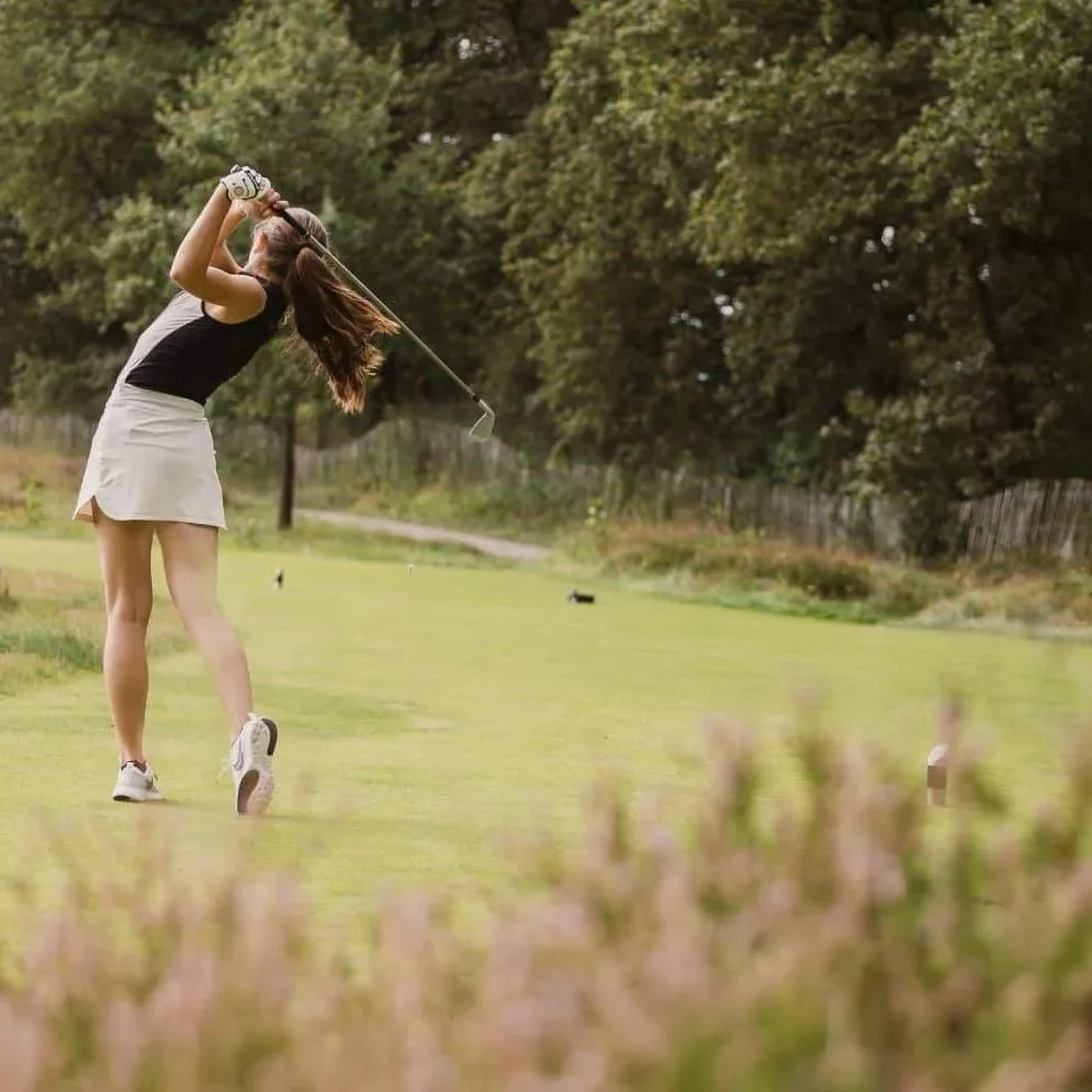 A woman playing golf on a grassy course, swinging a golf club with trees in the background.