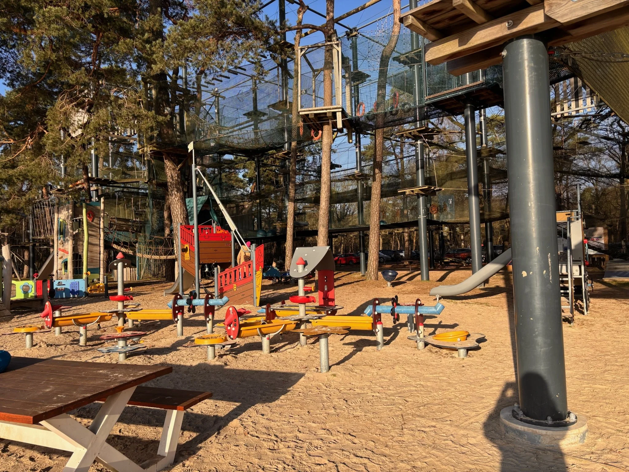 A playground featuring a multi-level treehouse structure connected by rope bridges and platforms, surrounded by trees and with various slides, climbing equipment, and colorful play panels on the sandy ground.