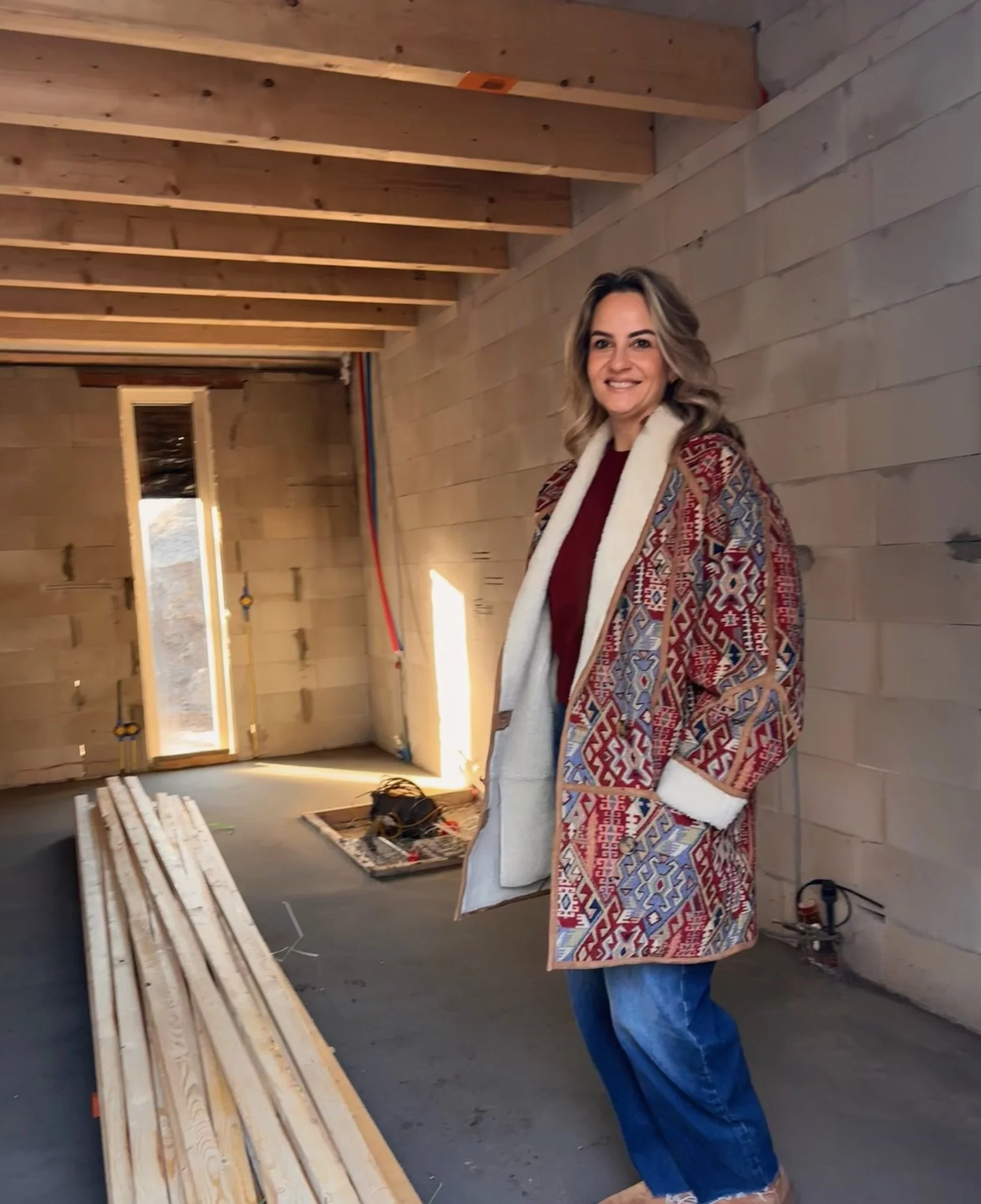 A woman smiling at a construction site, holding a framed piece of artwork with a geometric pattern. She is wearing a patterned coat and jeans. Wooden beams and construction materials are visible in the background.