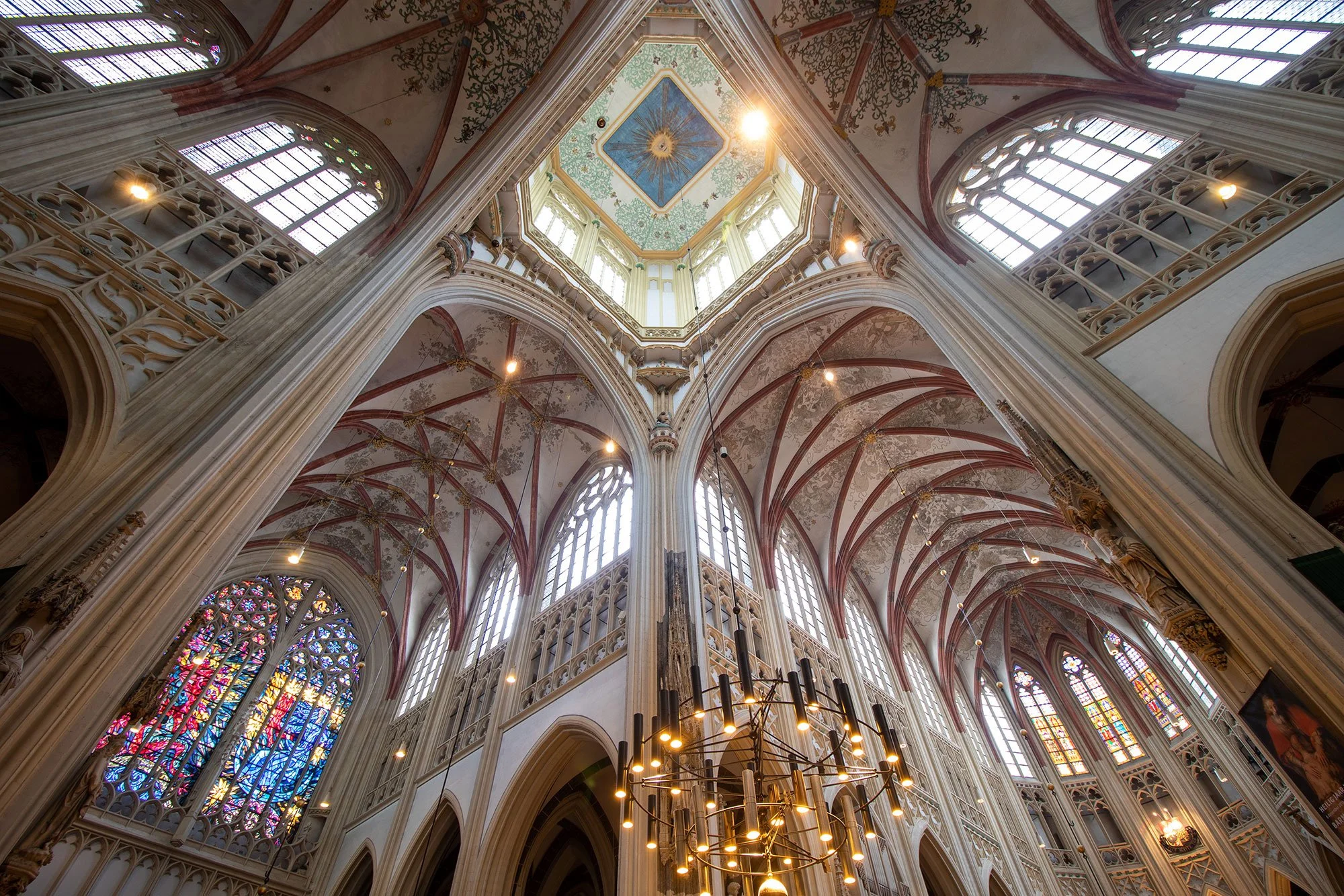 Interior view of a Gothic cathedral with tall stained glass windows, ornate vaulted ceilings, and a painted ceiling with a sun and rays design, illuminated by hanging lights.