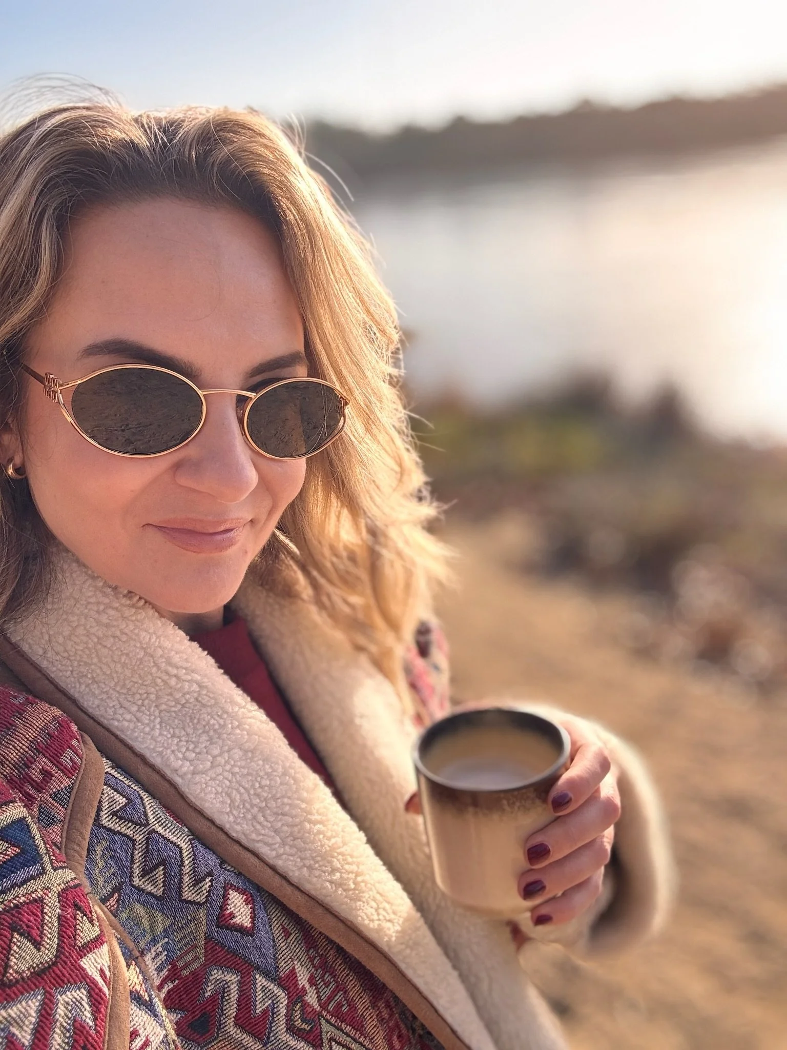 Woman with blonde hair, sunglasses, and a patterned jacket holding a cup of coffee outdoors near a body of water.