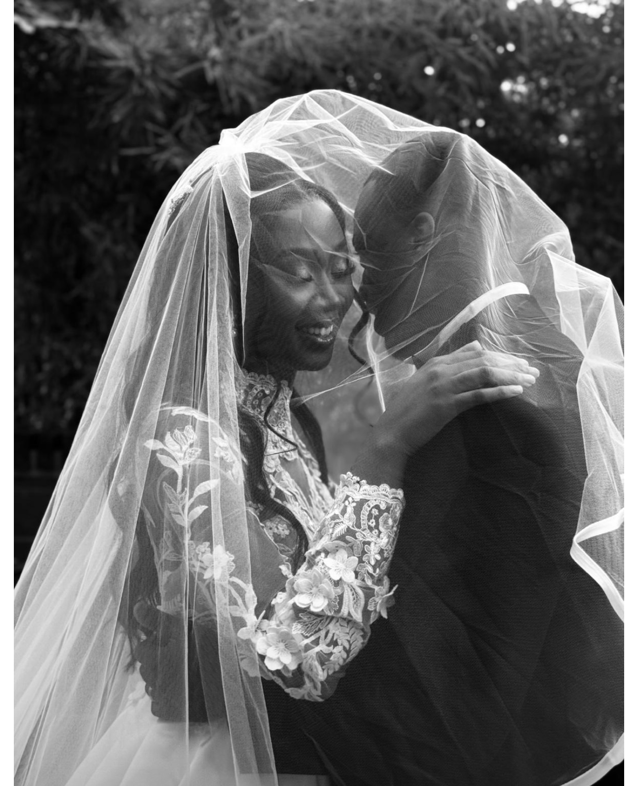 Black and white photo of a bride and groom under a wedding veil, embracing and sharing a moment of closeness outdoors.
