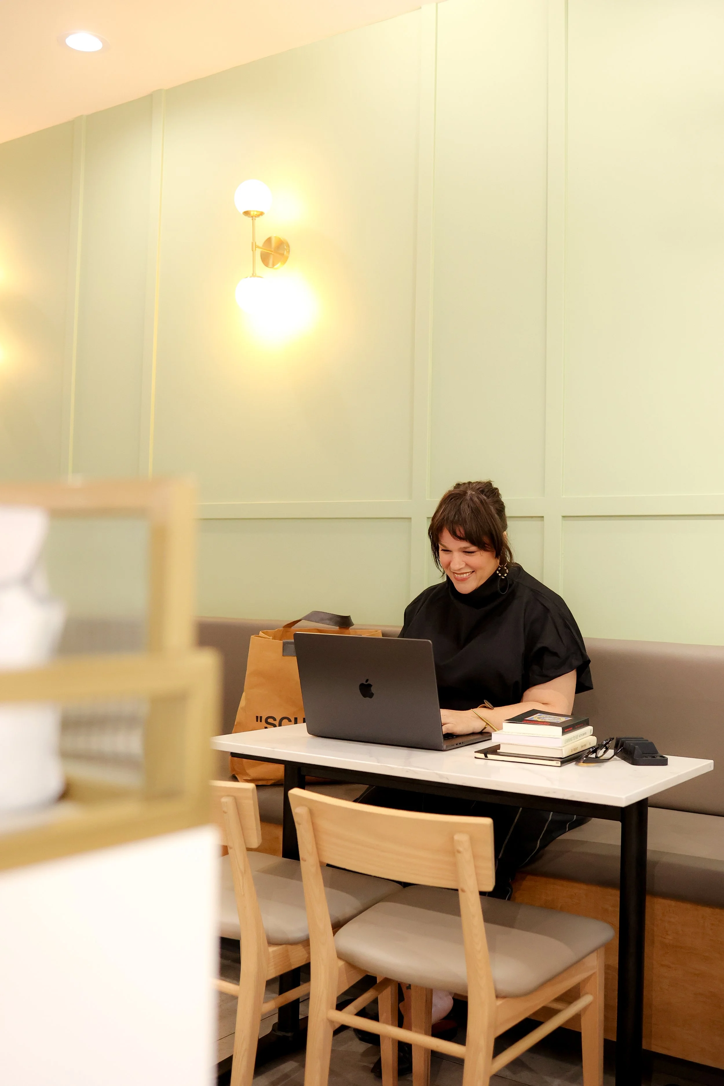 A woman smiling while working on a laptop at a small table in a modern cafe.