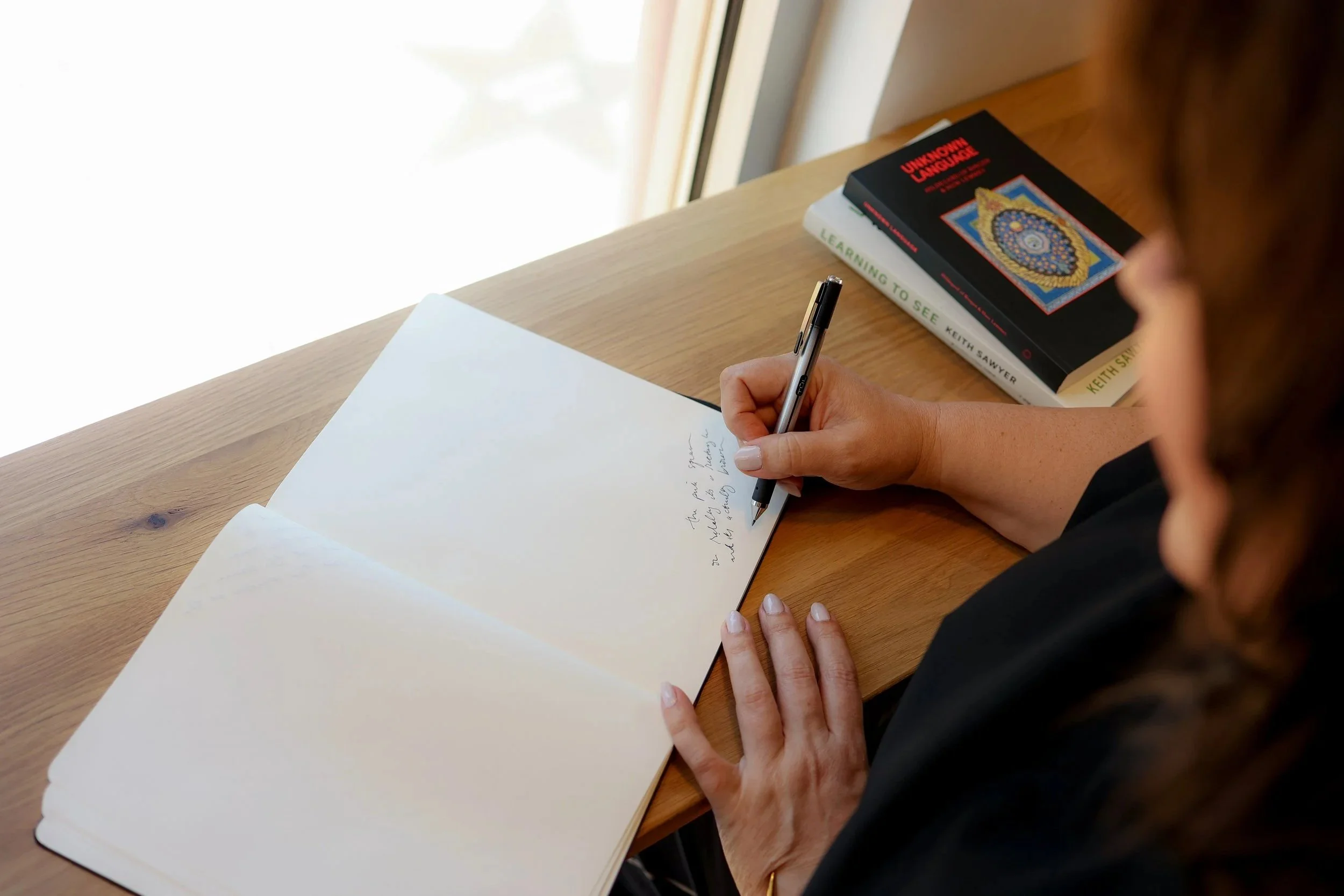 A person writing in a notebook at a wooden desk near a window, with two books stacked on the desk.