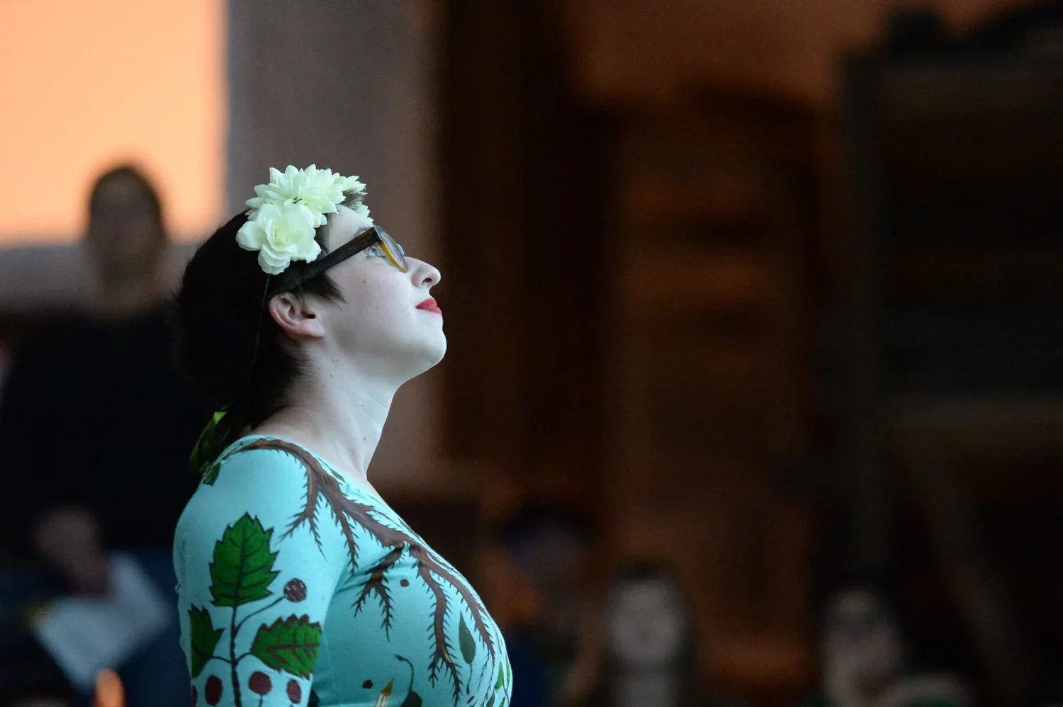 Comics artist Marnie Galloway, wearing glasses and a floral headband and dressed in a colorful costume, is looking upward with a serene expression during a performance in a dimly lit room with blurred audience members in the background.