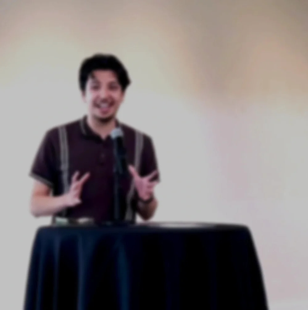 A young man with dark hair smiling at a speaking event, standing behind a black table with a microphone on it, in front of a plain wall.