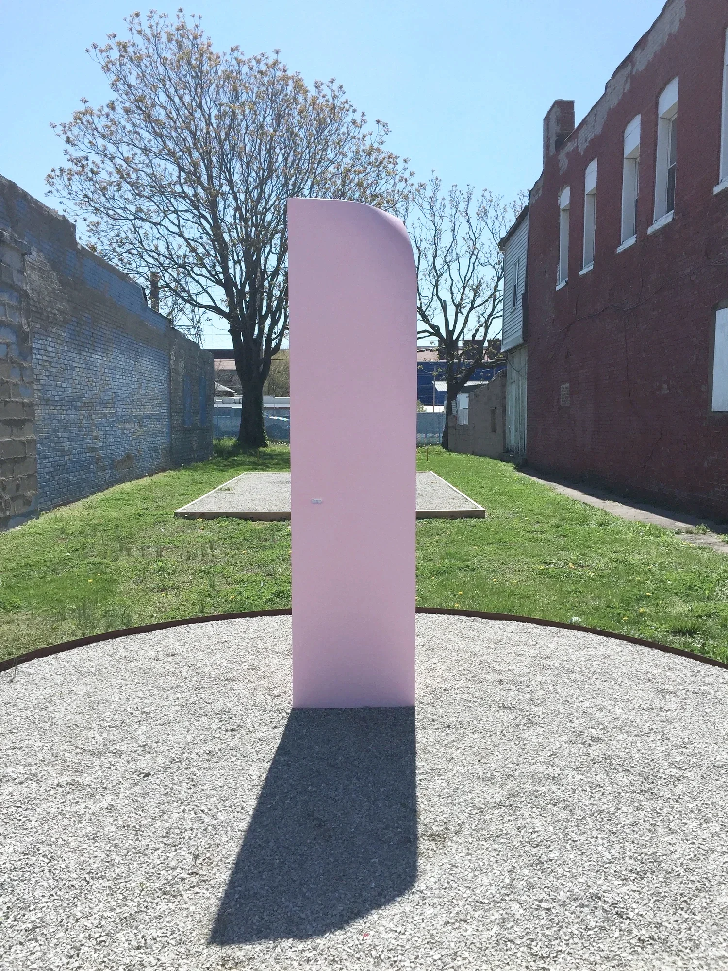 A large pink vertical sculpture by Cato Ouyang positioned on a gravel surface, casting a shadow on the ground, with trees and brick buildings in the background under a clear, sunny sky.