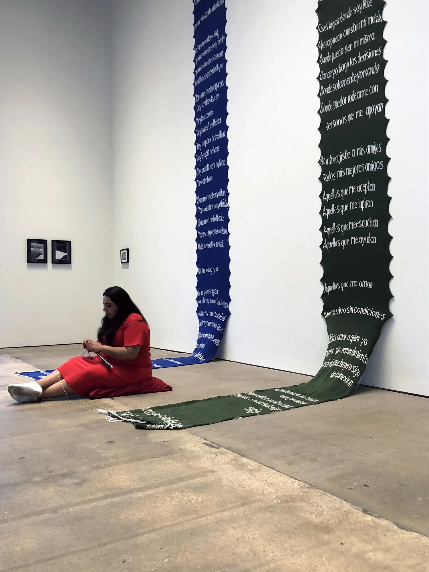 A student enacting a thesis performance sitting on the floor of a gallery unravelling yarn with her hands, surrounded by tall knitted banners with text hanging on a white wall.