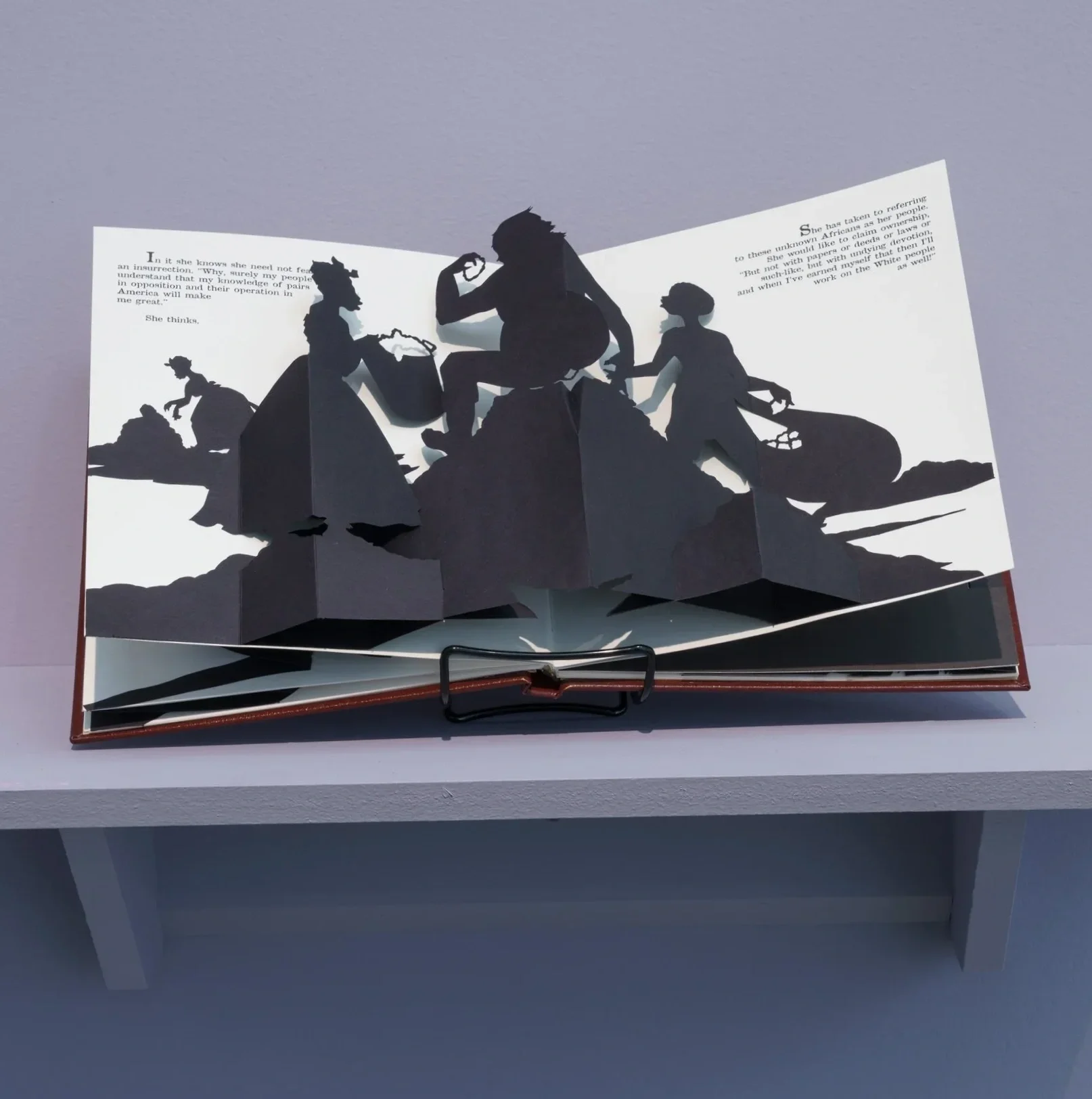 Open artists book by Kara Walker with black paper cutout silhouettes of women on a mountain, with text on the pages.