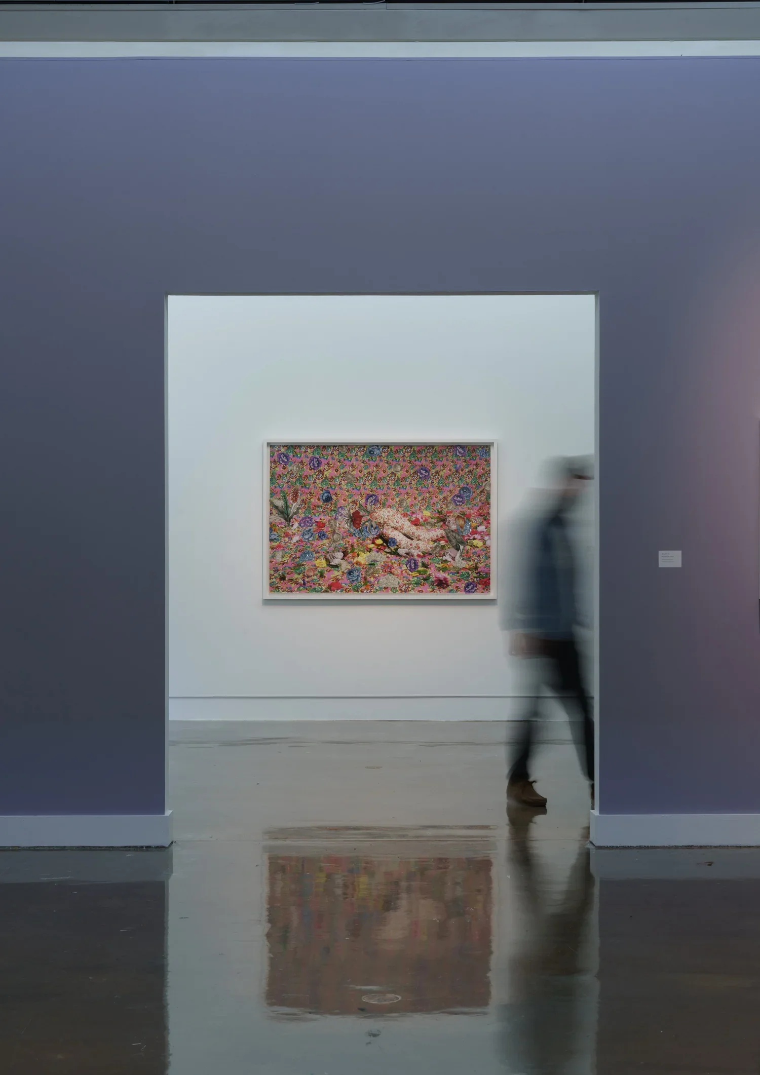 A person walking through an art gallery with a colorful, floral-themed painting by Ebony G. Patterson on the wall behind them, reflected on the polished wooden floor.