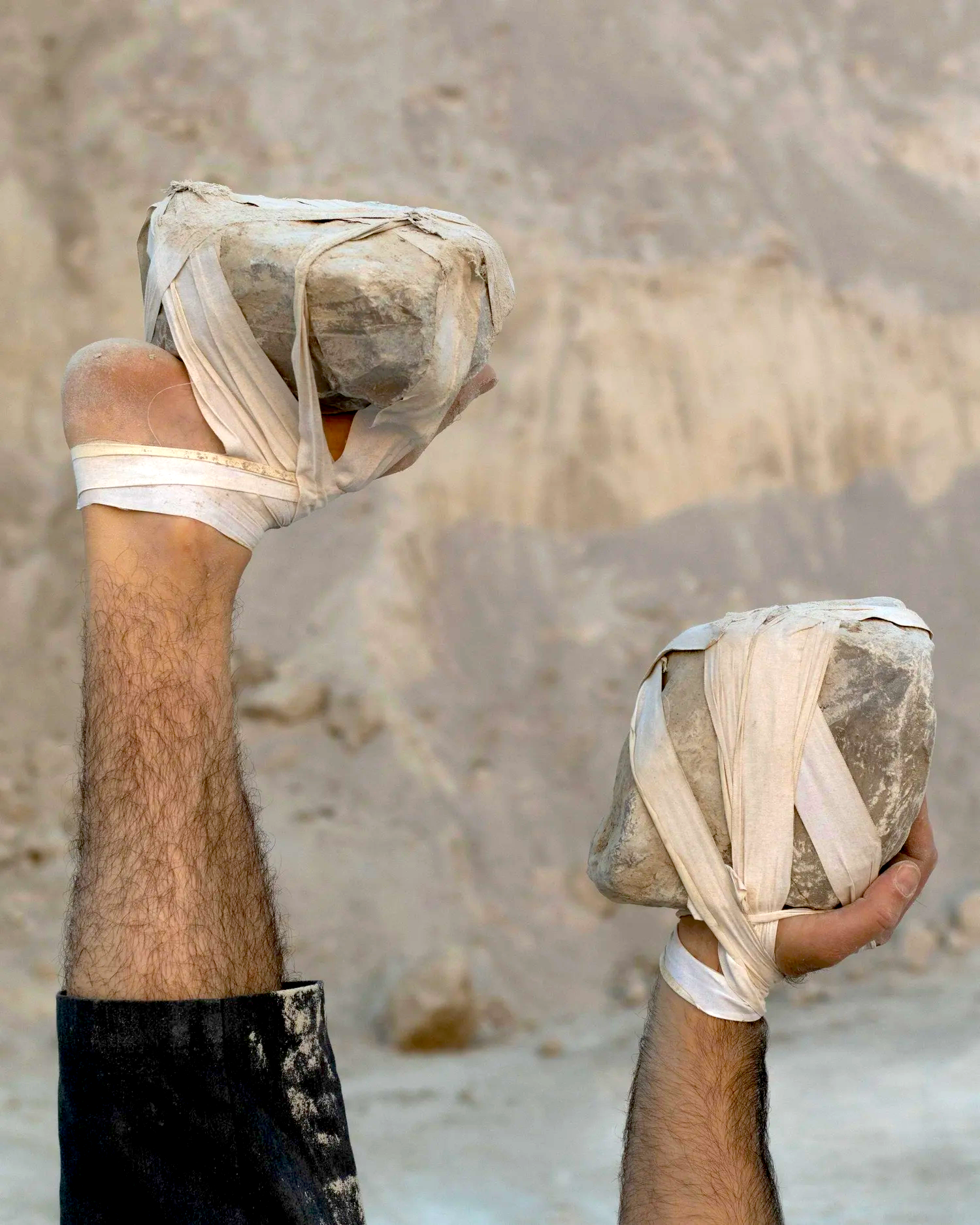 Two muscular arms and hands with hairy forearms holding large rocks wrapped in bandages or cloth against a rocky, desert-like background from an outdoor performance by Ben Gould.