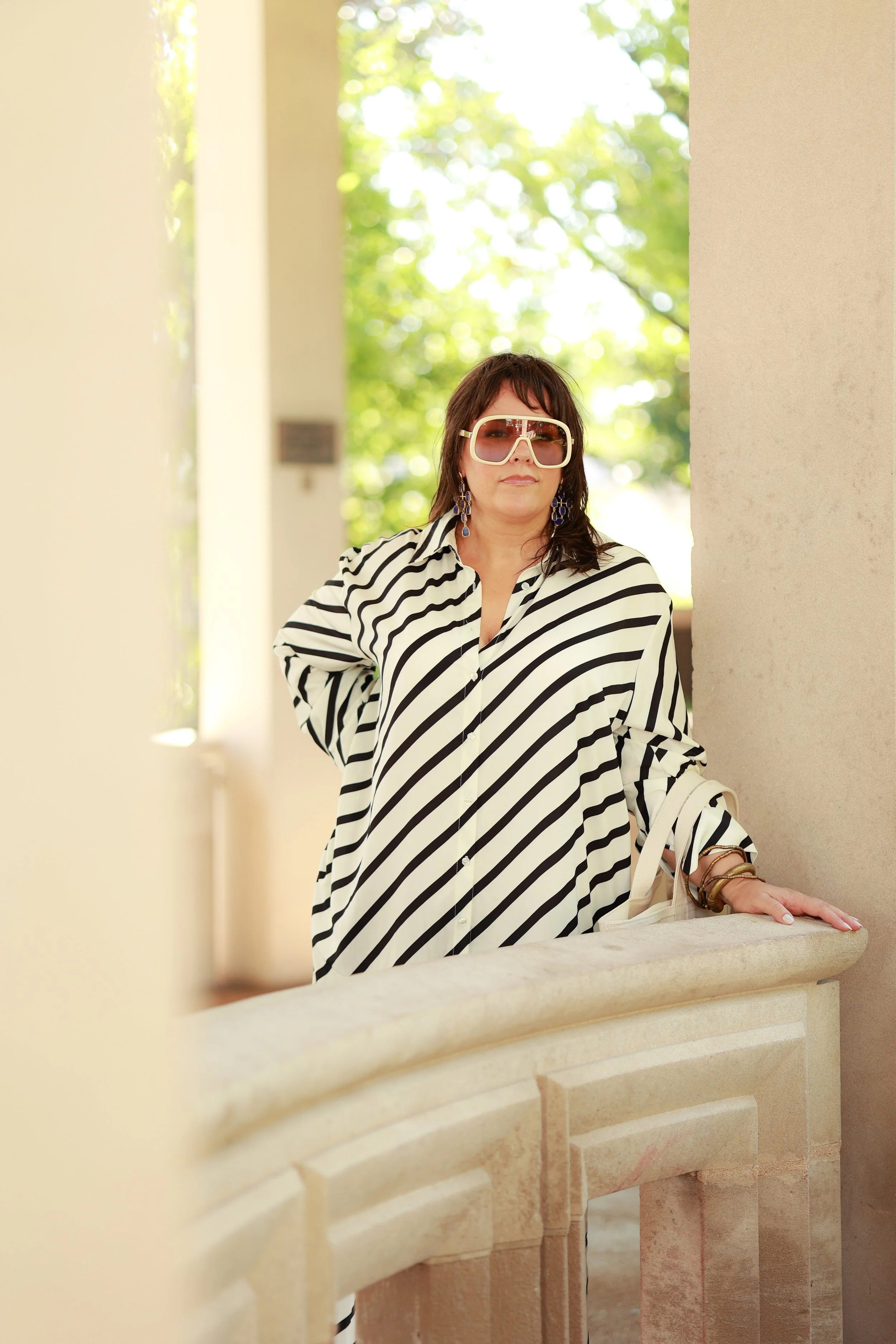 Woman wearing large sunglasses, a black-and-white striped shirt, and earrings, leaning against a stone railing outdoors with green trees in the background.