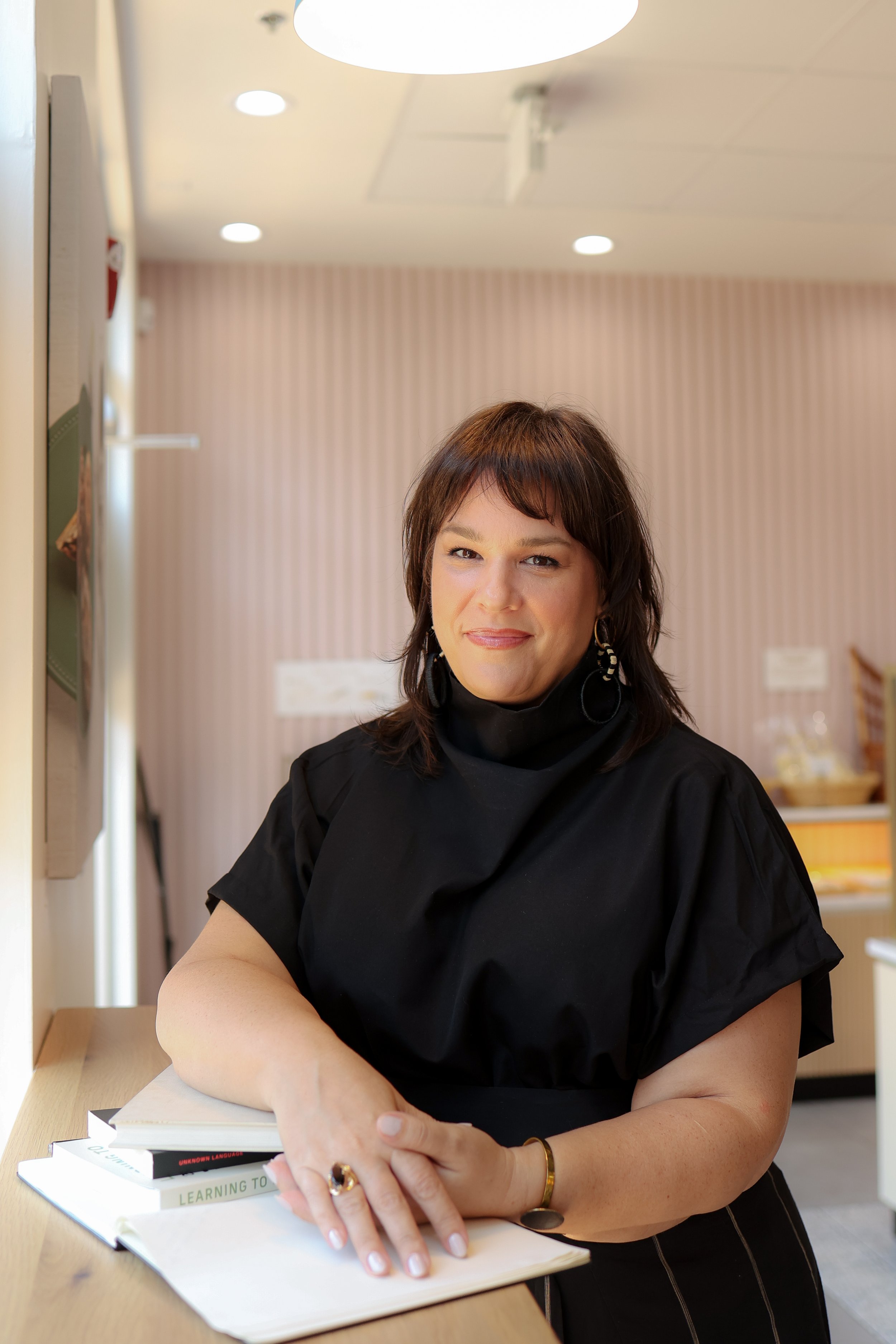 A woman with shoulder-length brown hair wearing a black top and earrings, smiling and leaning on a desk with books in a room with pink striped wallpaper and ceiling lights.