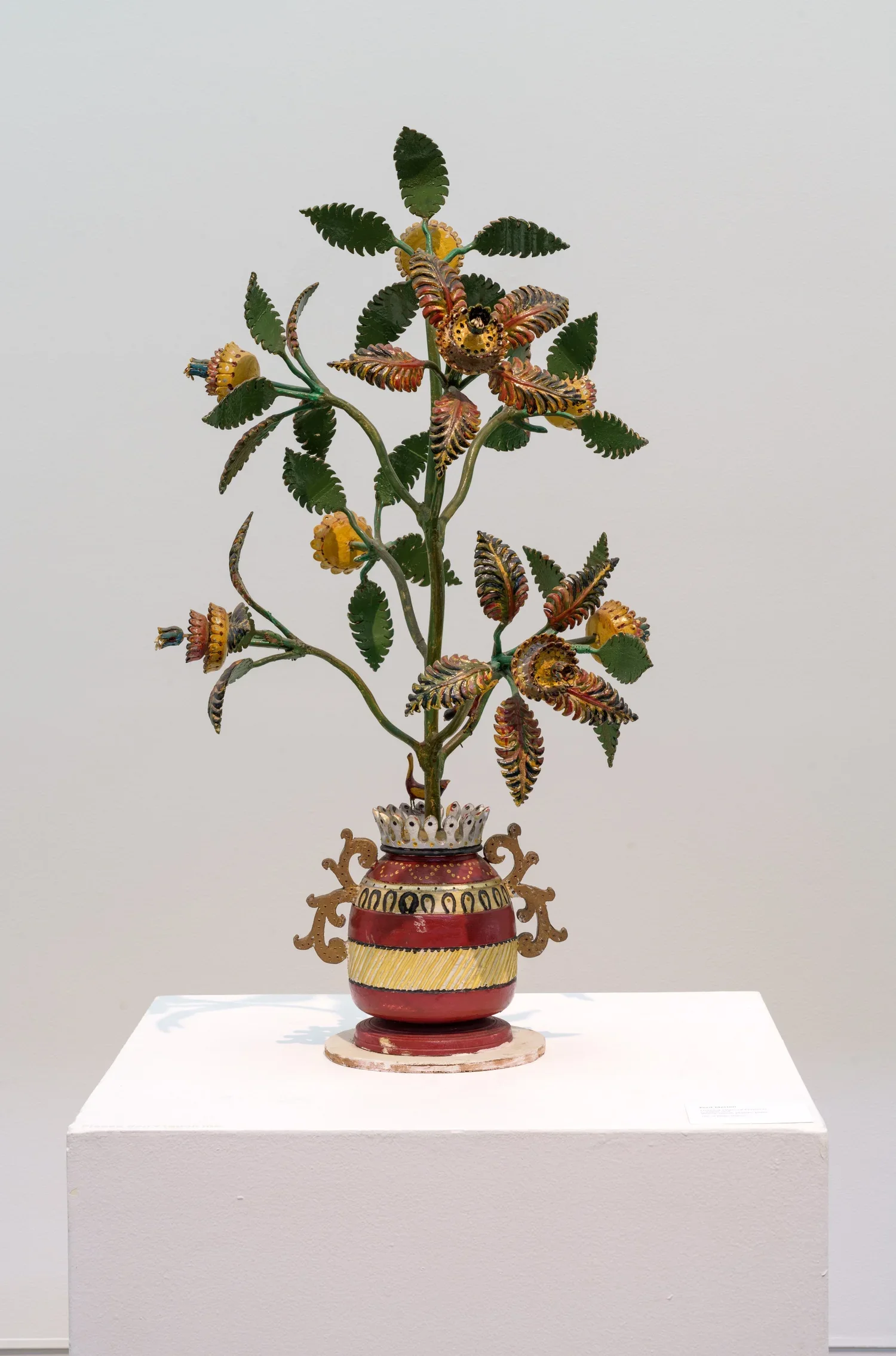 Colorful wooden sculpture by Ferd Metten of a plant with large leaves and curled flowers in a decorative vase, displayed on a white pedestal in a museum against a plain background.