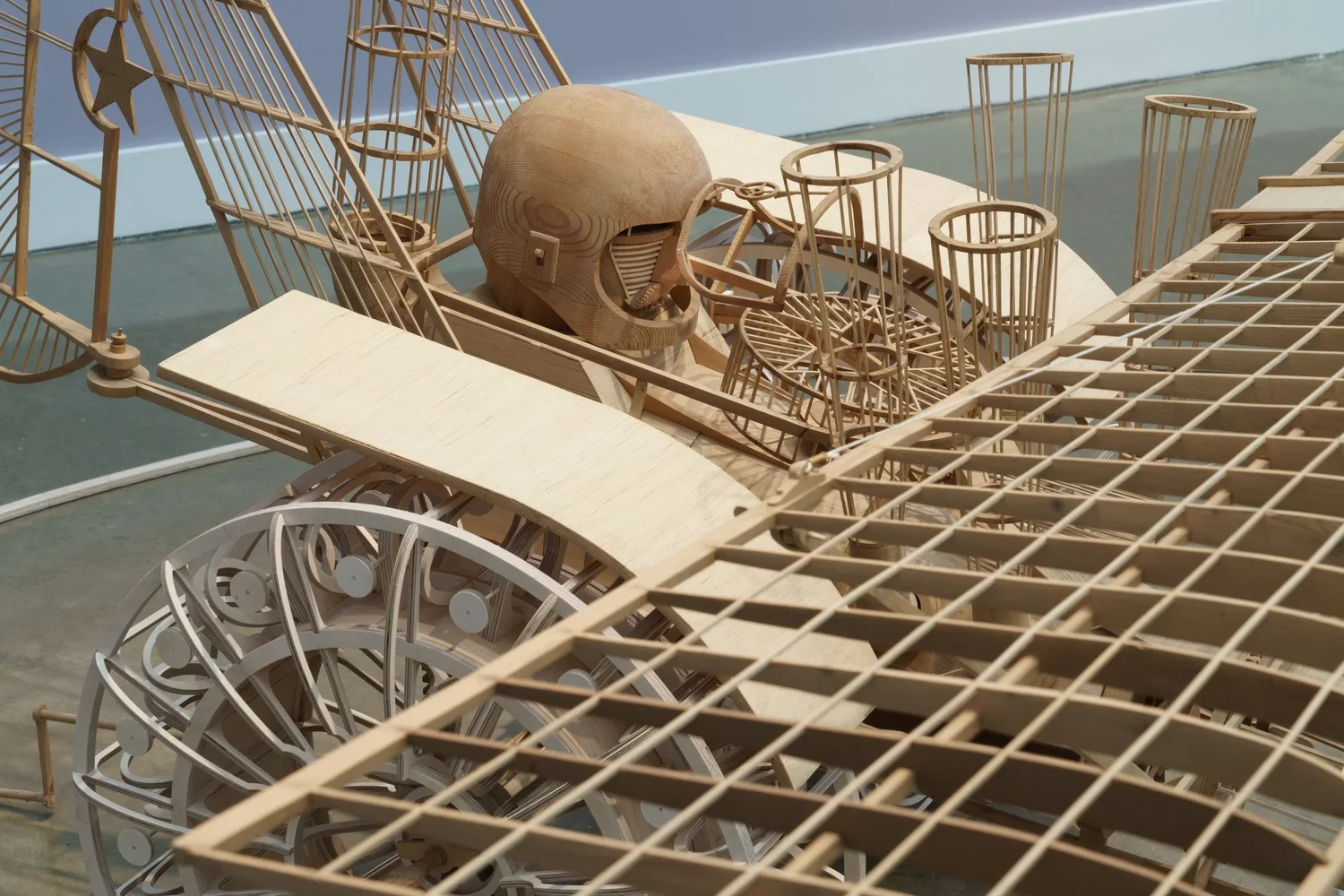 A detailed wooden sculpture by Carl Wilen of a futuristic vehicle, featuring a helmet-wearing driver with shutter shades in the cockpit, various cylindrical and complex structural elements, and a  wheel at the front.