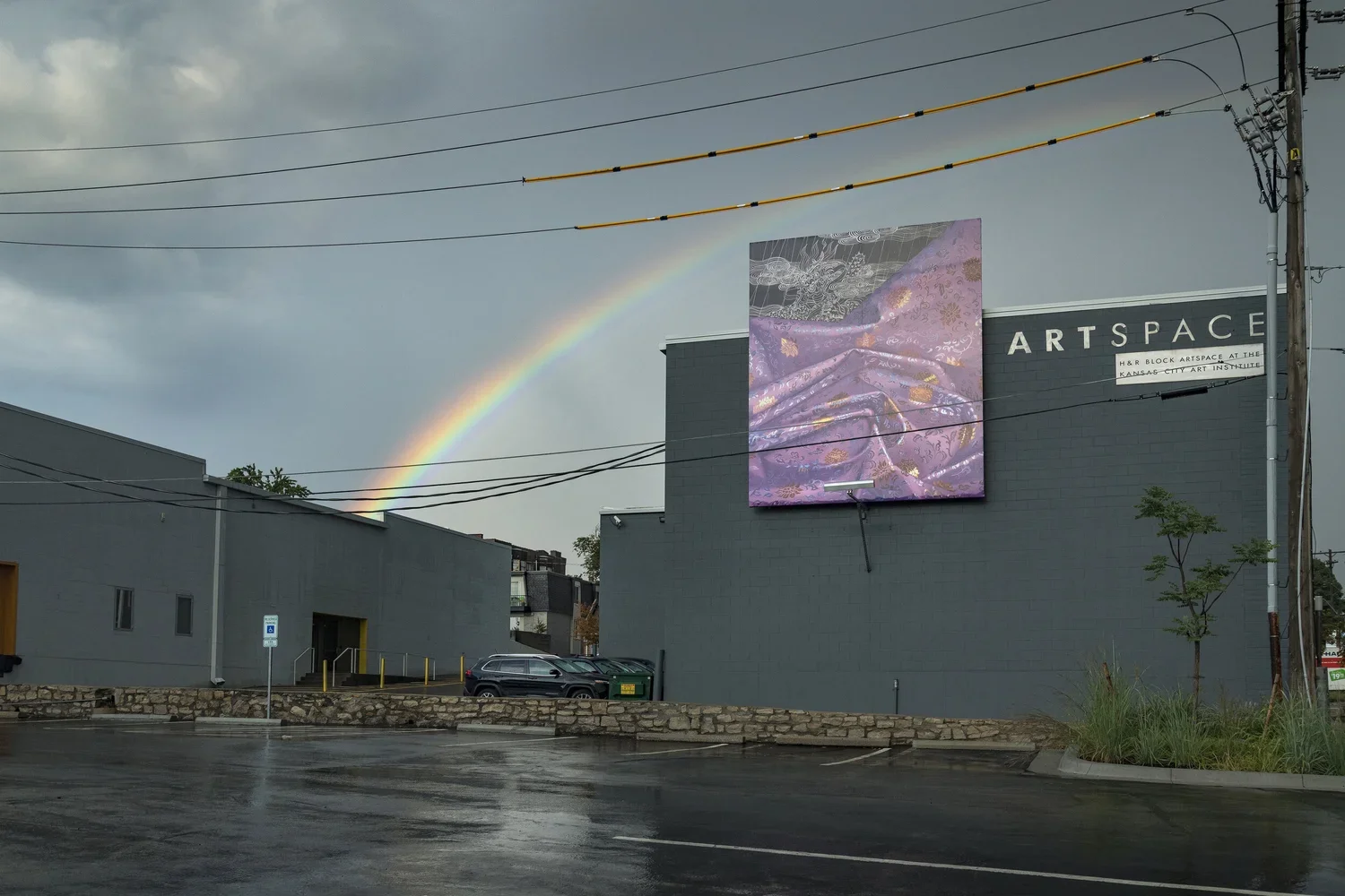 A purple artwork by Leeah Joo displayed on a billboard on a gray building. A rainbow appears in a cloudy sky above the building. The parking lot is wet, with a couple of cars parked near the building and a small tree planted in a landscaped area.