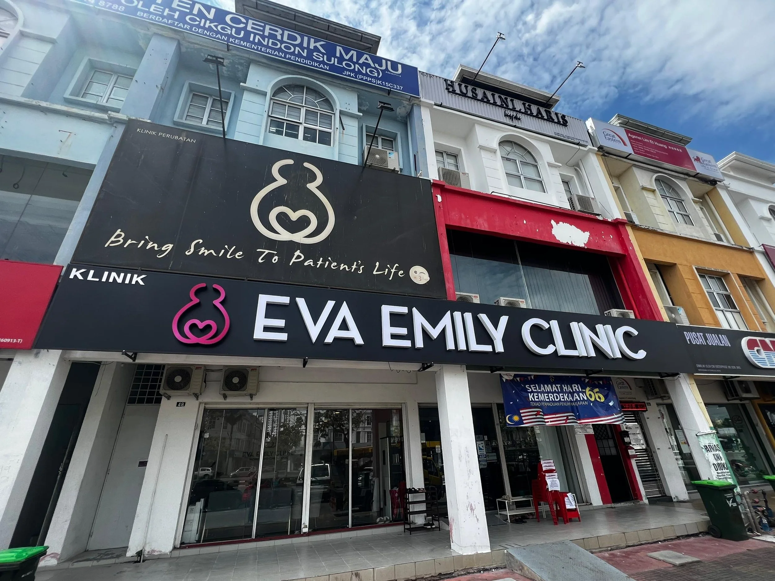 Facade of EVA Emily Clinic with black signage featuring white and pink logo and text, multiple floors, surrounding buildings with various signs and windows, cloudy sky, and a glass door entrance.