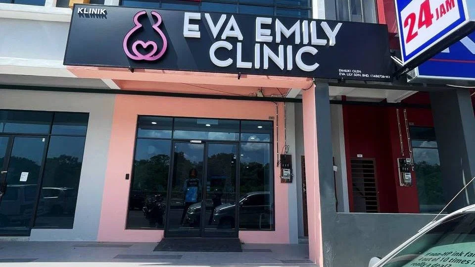 Exterior view of Eva Emily Clinic, a medical clinic with a black signboard displaying a pink rabbit outline and the clinic's name. The entrance has glass doors and tinted windows, with reflections of parked cars and trees outside.