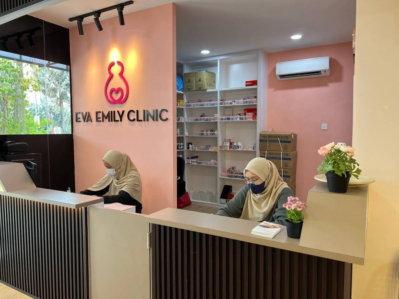 Reception area of Eva Emily Clinic with two women wearing hijabs and masks, sitting at desks with flowers and stacks of papers, shelves with medication in the background, and a pink wall with the clinic's logo.